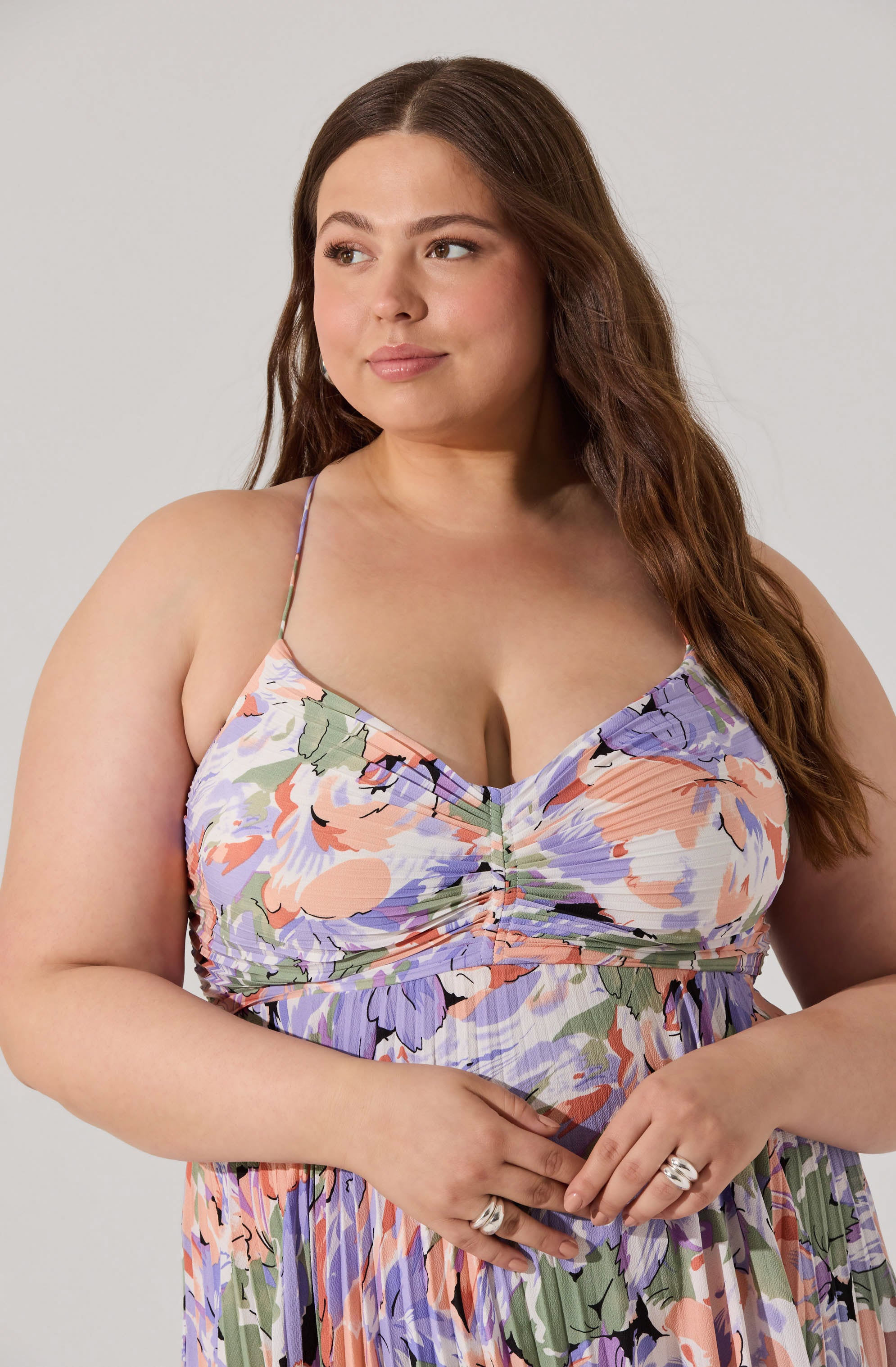 A woman wearing a colorful, floral-patterned dress with spaghetti straps. The dress features shades of purple, peach, green, and white, with a gathered detail at the bust. She has long, wavy brown hair and is wearing rings on her fingers.