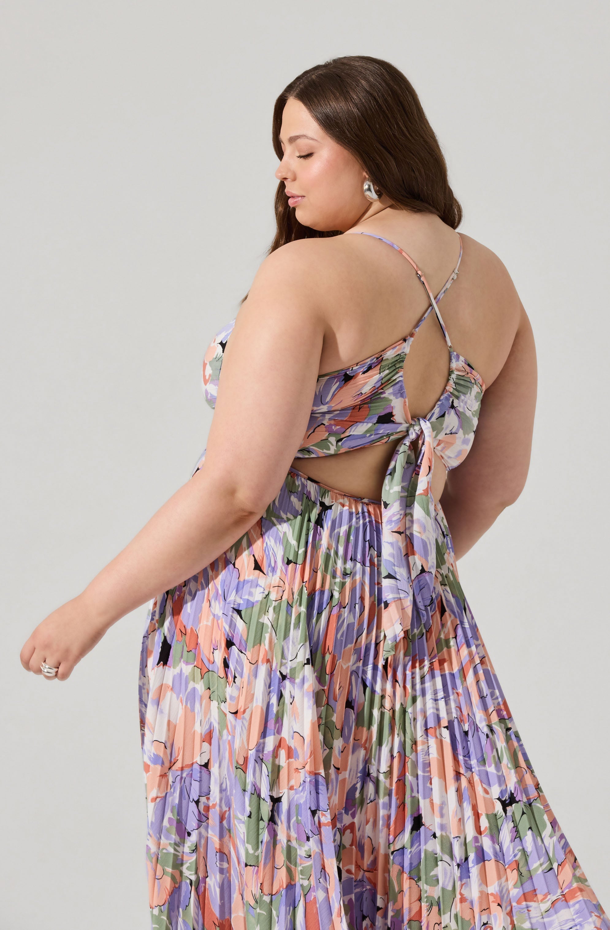 A woman is wearing a sleeveless, floral dress with thin spaghetti straps. The dress features a cut-out back with a bow tie detail, and the fabric has a colorful pattern with shades of purple, green, peach, and black. The skirt is long and flowing, with a gathered, pleated design.