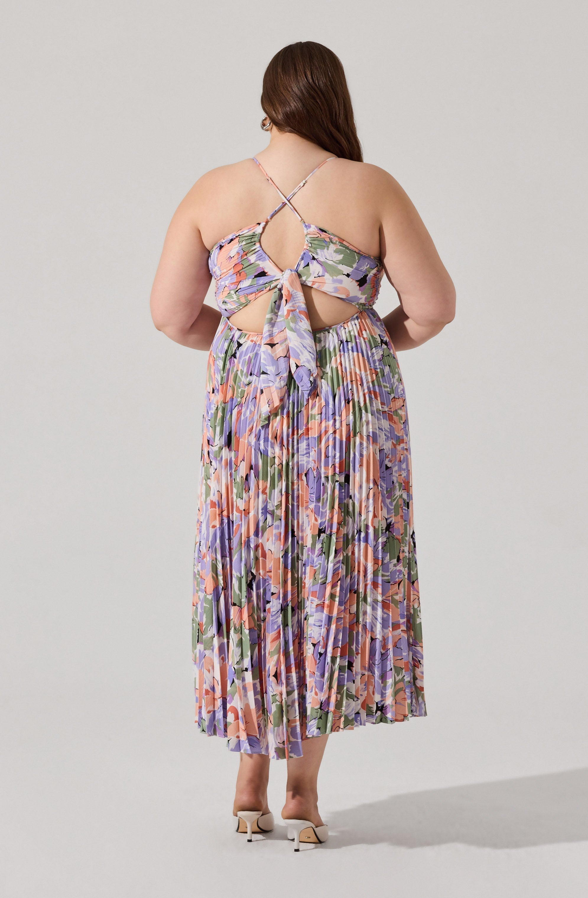 A woman wearing a colorful, floral-patterned dress with a pleated midi skirt. The top part of the dress has thin, crisscrossing spaghetti straps and a tie at the back, creating a cut-out design above the waist. The dress features soft shades of pink, purple, green, and orange. The woman is standing with her back to the camera, showcasing the back details of the dress, and is wearing white heeled shoes.