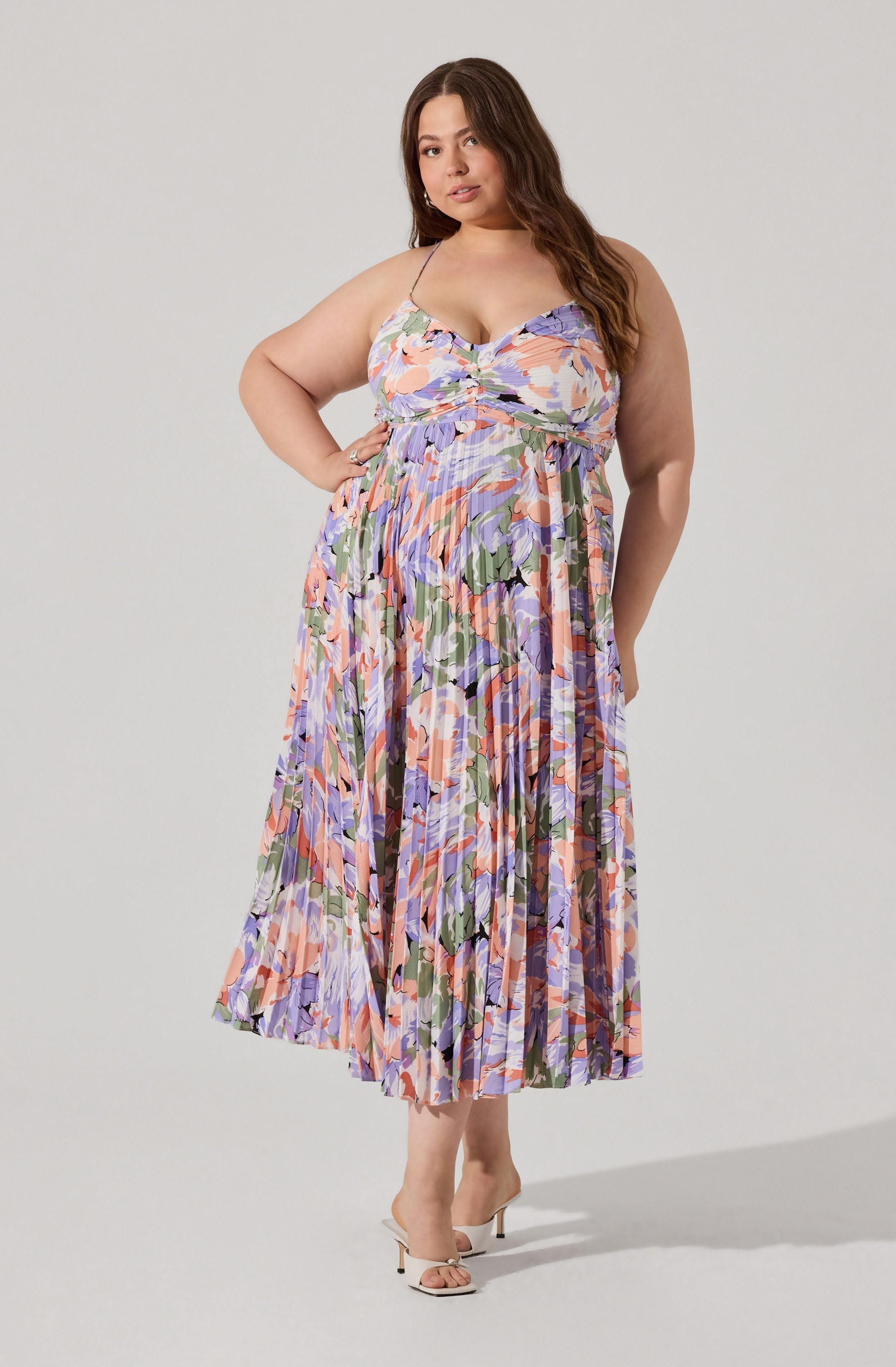 A woman is wearing a sleeveless, flowy dress with thin spaghetti straps. The dress features a colorful abstract floral pattern with shades of purple, pink, green, and black. It has a fitted bodice with a ruched detail in the center and a high waistline that flows into a pleated, ankle-length skirt. She is also wearing white, open-toe high heels with a small bow detail.