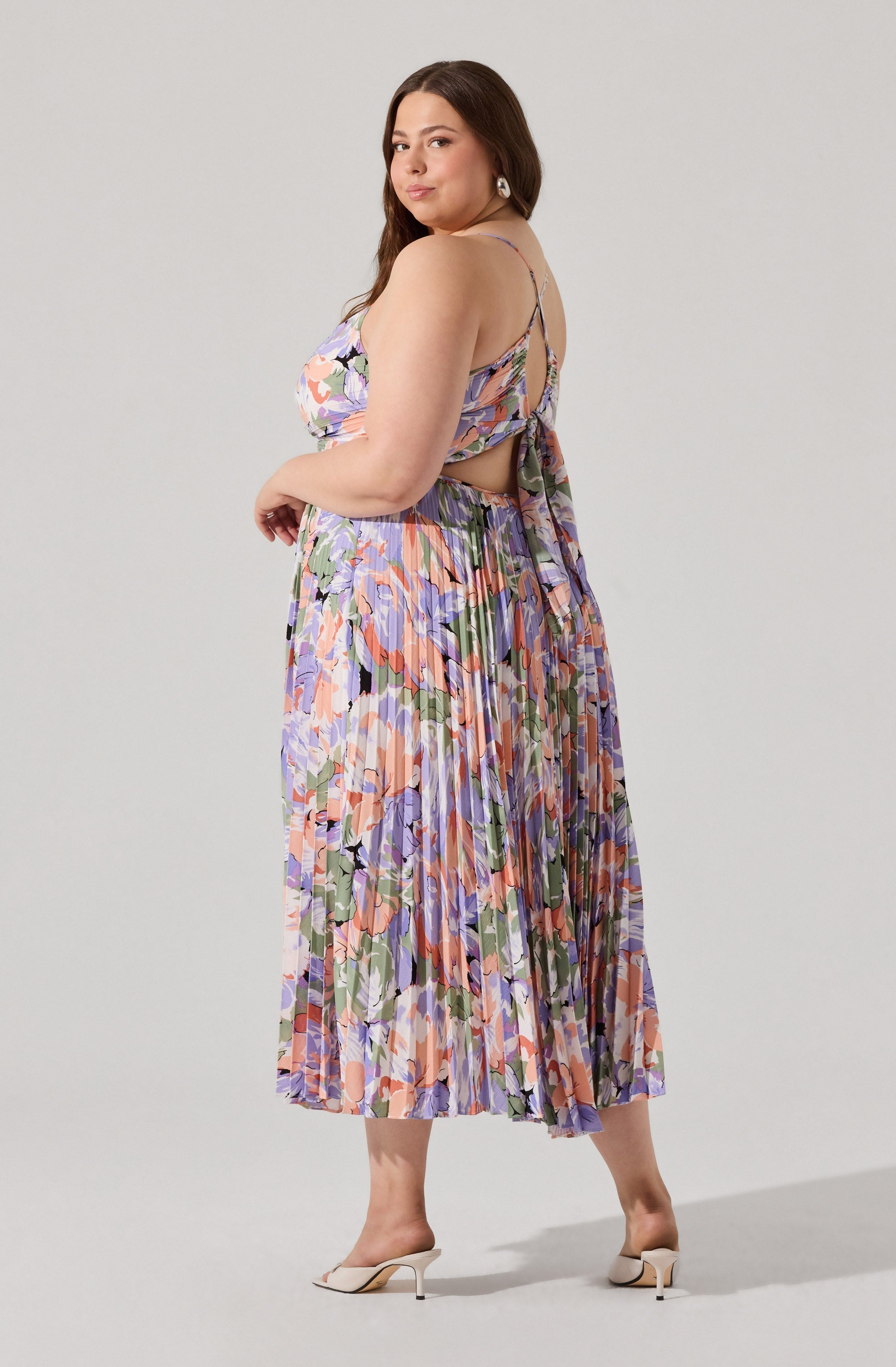A woman is wearing a sleeveless, floral-patterned dress with spaghetti straps and a cutout back, featuring a tie detail at the back. The dress has a pleated skirt with a multicolored floral print in shades of pink, purple, green, and black. She completes her outfit with pair of white high-heeled shoes.