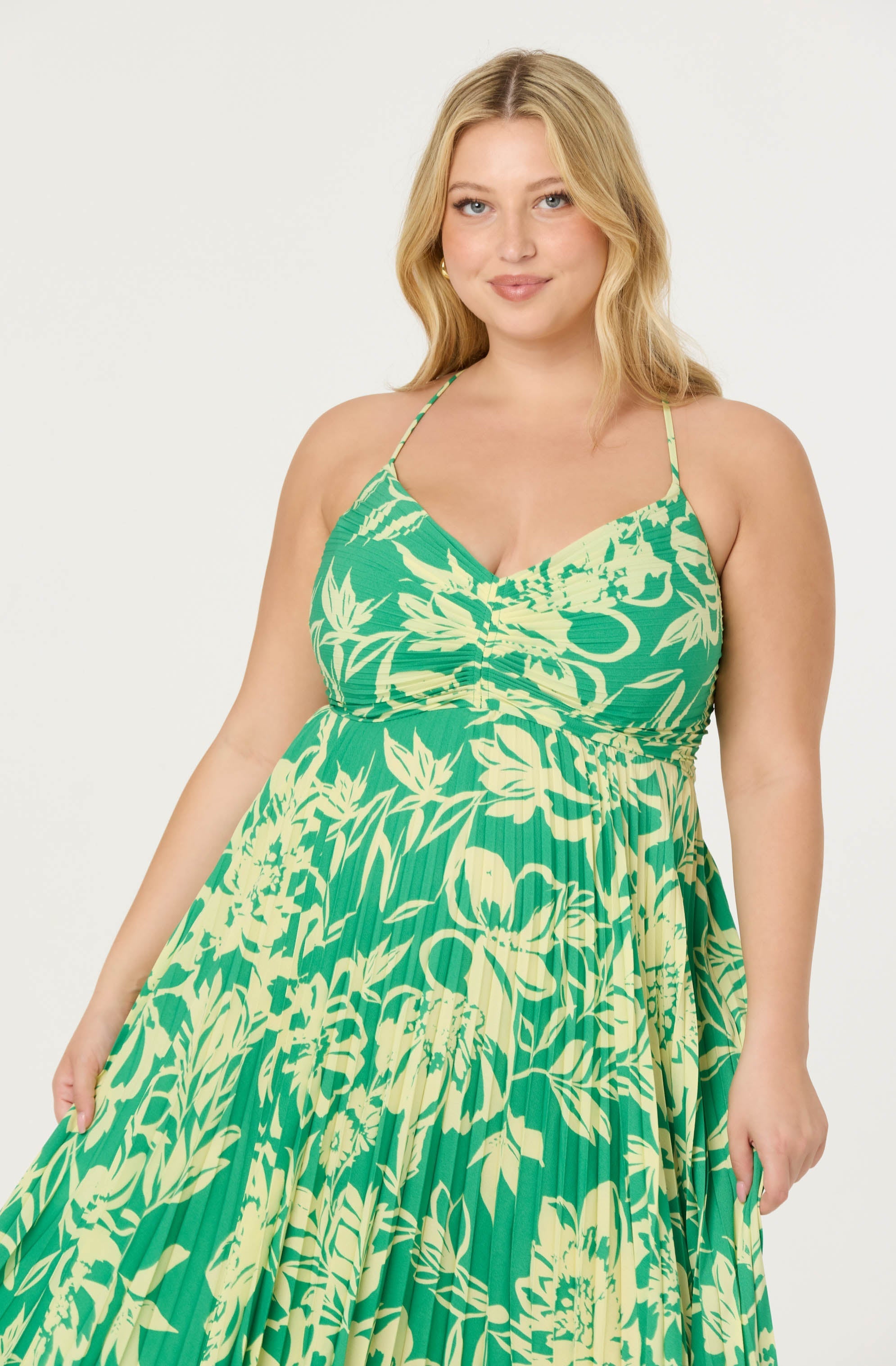 A woman is wearing a sleeveless, green dress with a yellow floral and foliage pattern. The dress features thin, adjustable spaghetti straps and a gathered detail at the bust. The fabric has a pleated texture throughout.