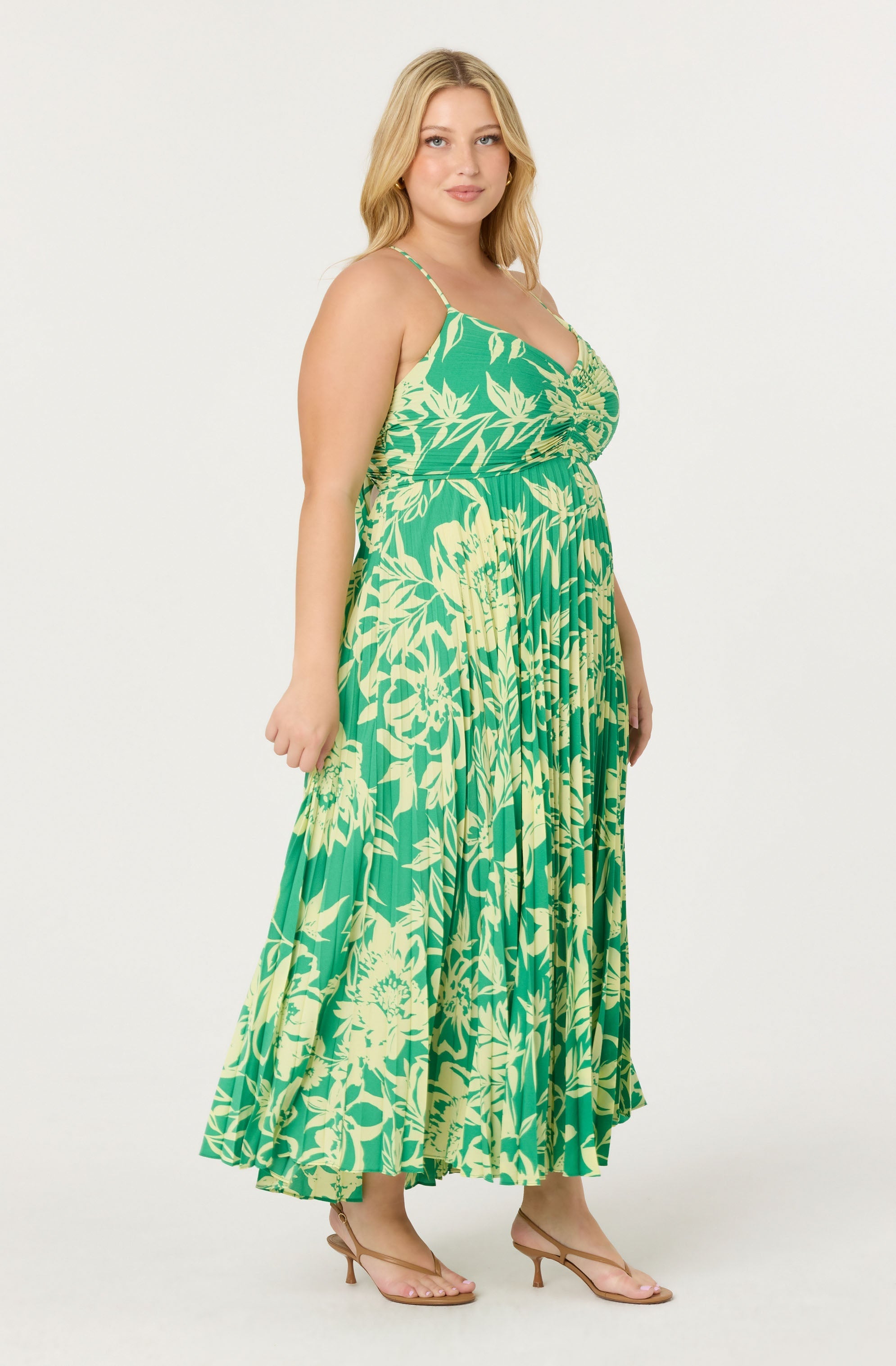 A woman is wearing a sleeveless, green maxi dress with a yellow floral and leaf pattern. The dress has thin spaghetti straps, a fitted bodice, and a flowing, pleated skirt. She is also wearing nude open-toe high heels.