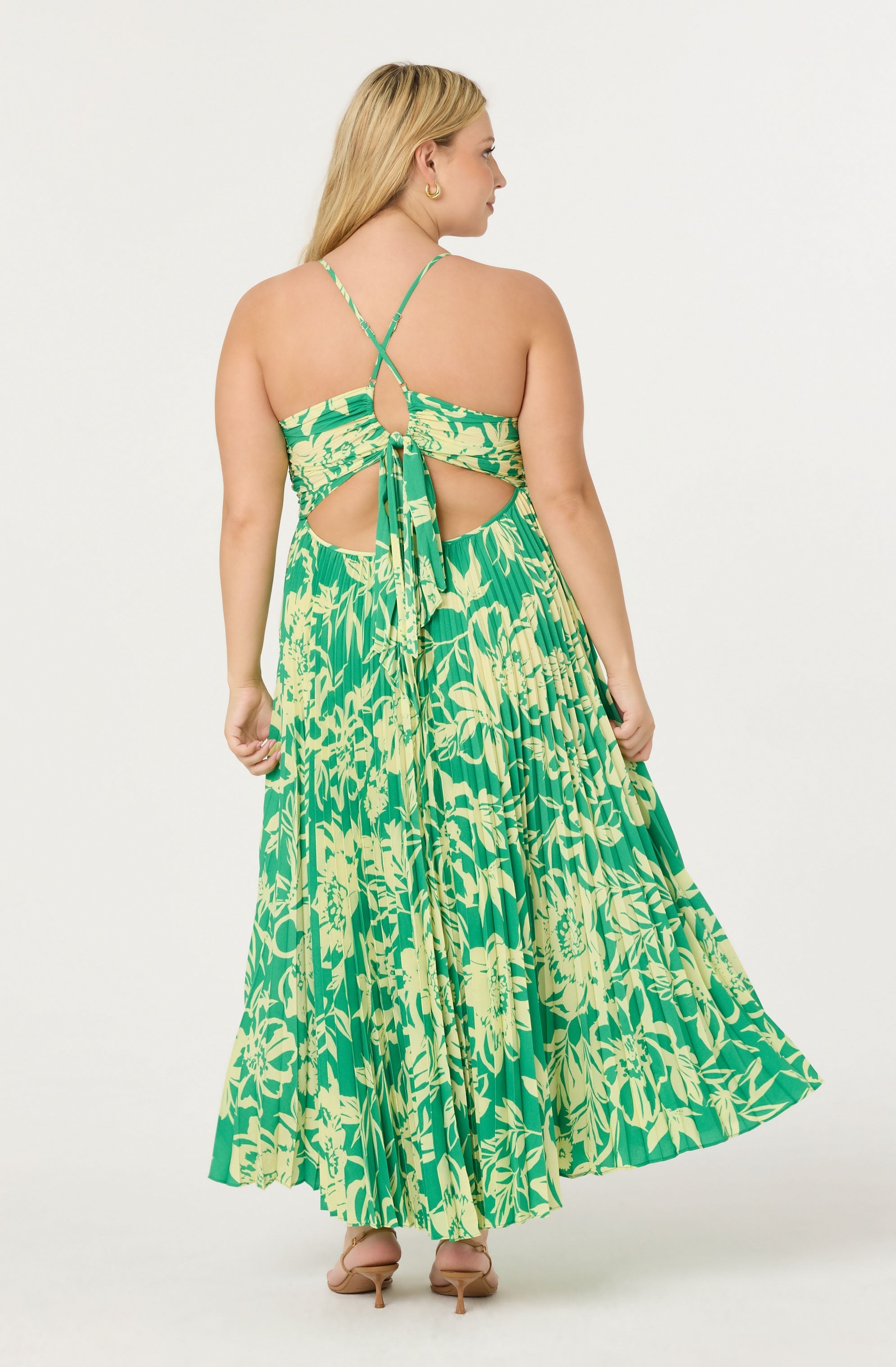 A woman is wearing a sleeveless, green and cream floral print dress with cutouts at the back. The dress features thin, crisscrossing shoulder straps that tie into a bow at the center of her back. The skirt is long, flowing, and pleated, with a vibrant floral pattern. She is also wearing beige high-heeled shoes.
