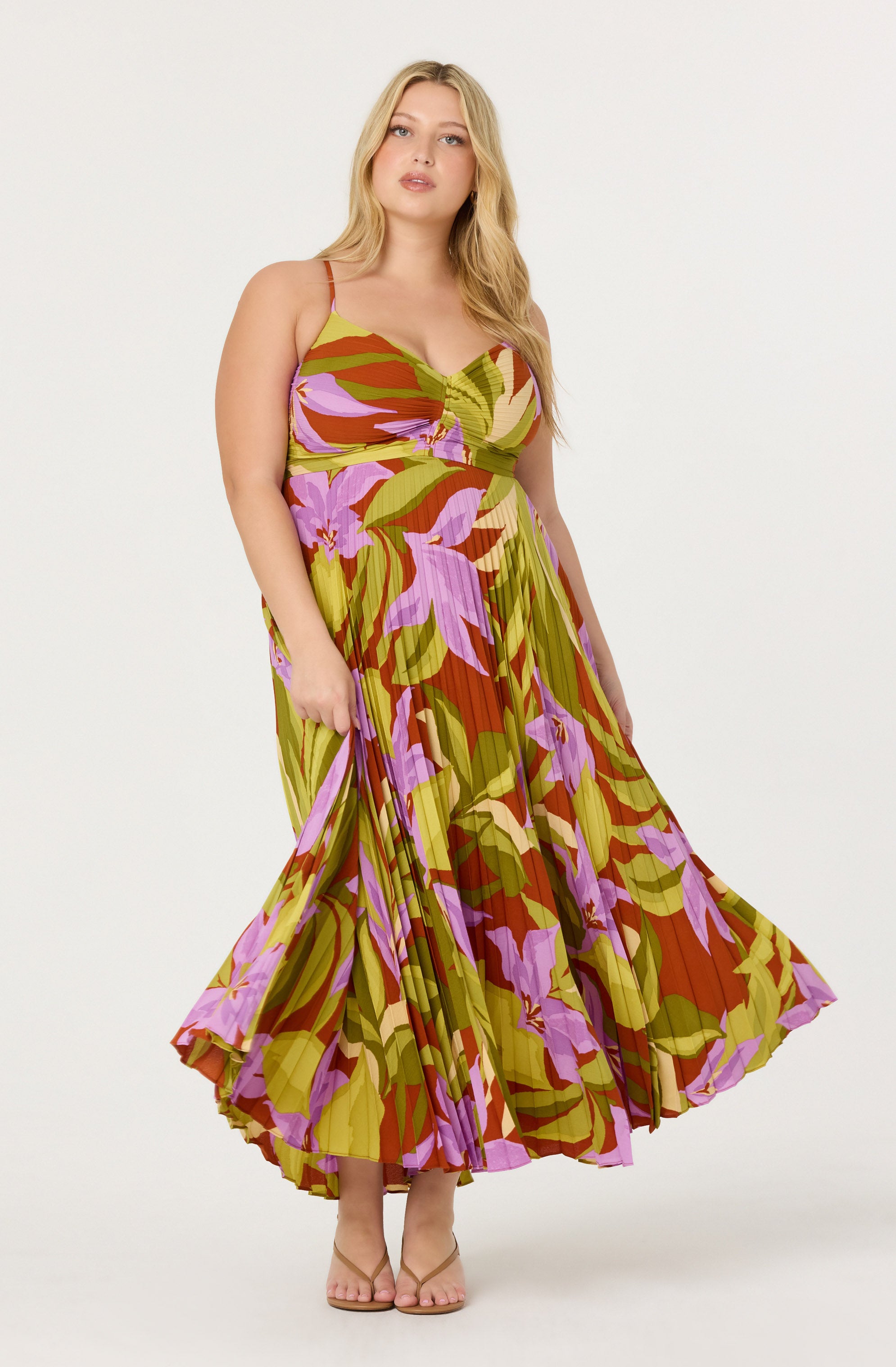 A woman wearing a sleeveless, flowy maxi dress with thin spaghetti straps. The dress features a vibrant floral pattern with large purple, pink, and green leaves on a background of red and yellow hues. The fabric is pleated, creating a textured and flowing appearance. She is standing against a plain, light-colored background, and her feet are in open-toed, nude-colored sandals.