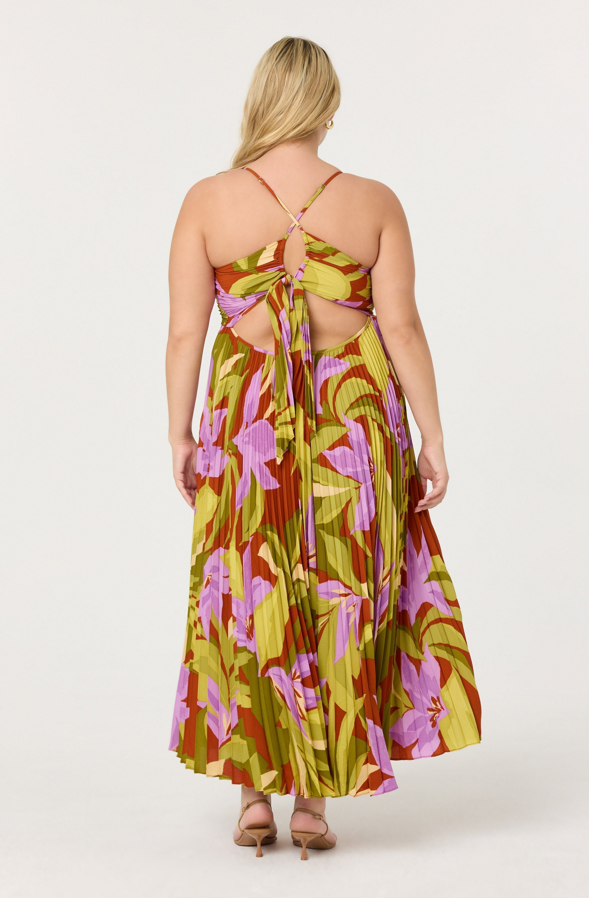 A woman is wearing a colorful, sleeveless, floral-printed dress with thin spaghetti straps and a back tie. The dress has a halter-style top with cutout details on the sides and a flowing, pleated skirt that reaches the floor. The fabric features a pattern of large pink and purple flowers with green and brown leaves on a yellow background. She is also wearing nude-colored heels.
