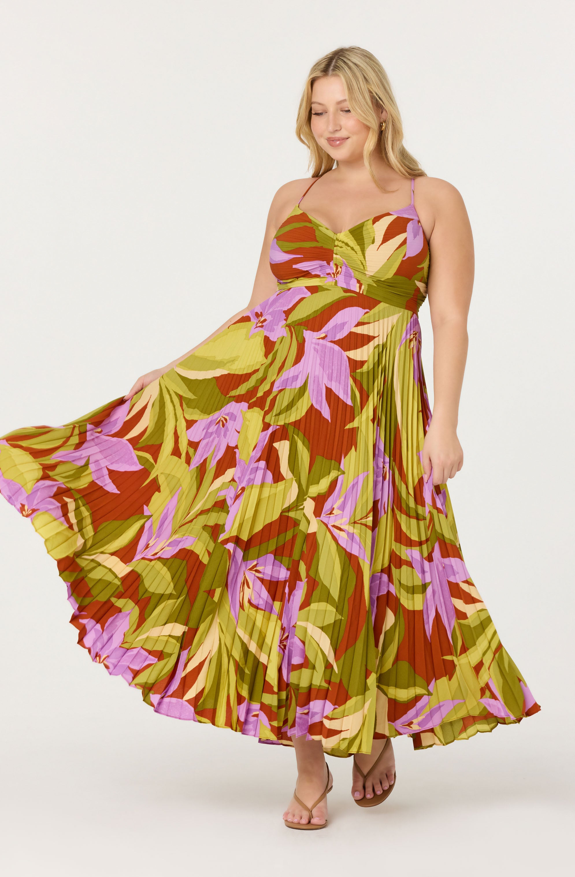 A woman is wearing a sleeveless, ankle-length dress with thin straps. The dress features a vibrant, colorful botanical print with large leaves and flowers in shades of green, purple, pink, and reddish-brown. The fabric appears lightweight and flowy, with a pleated design that adds texture and movement to the dress. The woman is holding the skirt out to the side, showcasing its fullness and pattern.