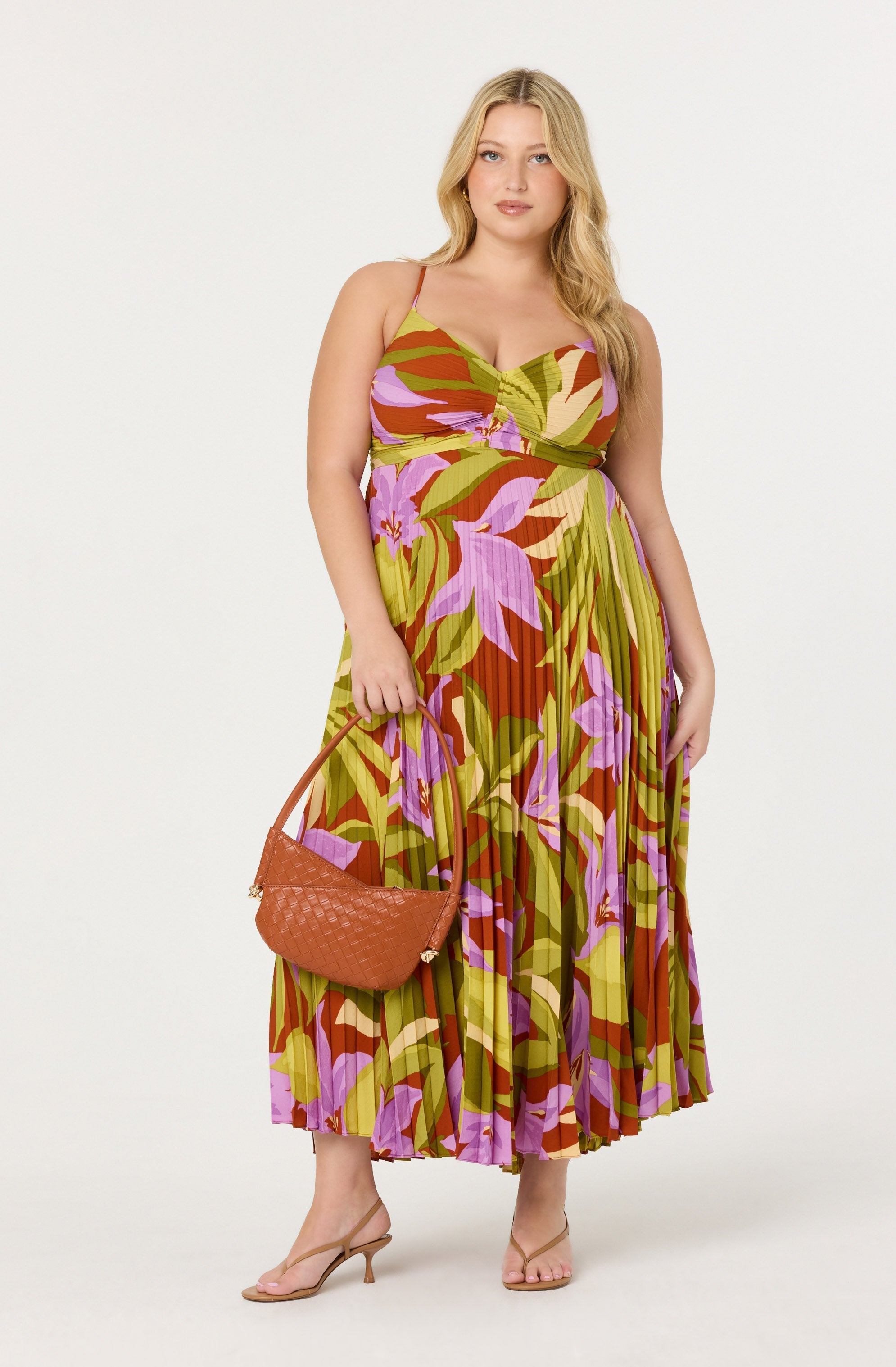 A woman is wearing a sleeveless, ankle-length dress with thin spaghetti straps. The dress features a vibrant, colorful floral pattern with shades of green, purple, pink, and brown. She is holding a medium-sized, tan woven handbag in her right hand and is wearing nude, strappy high-heeled sandals.