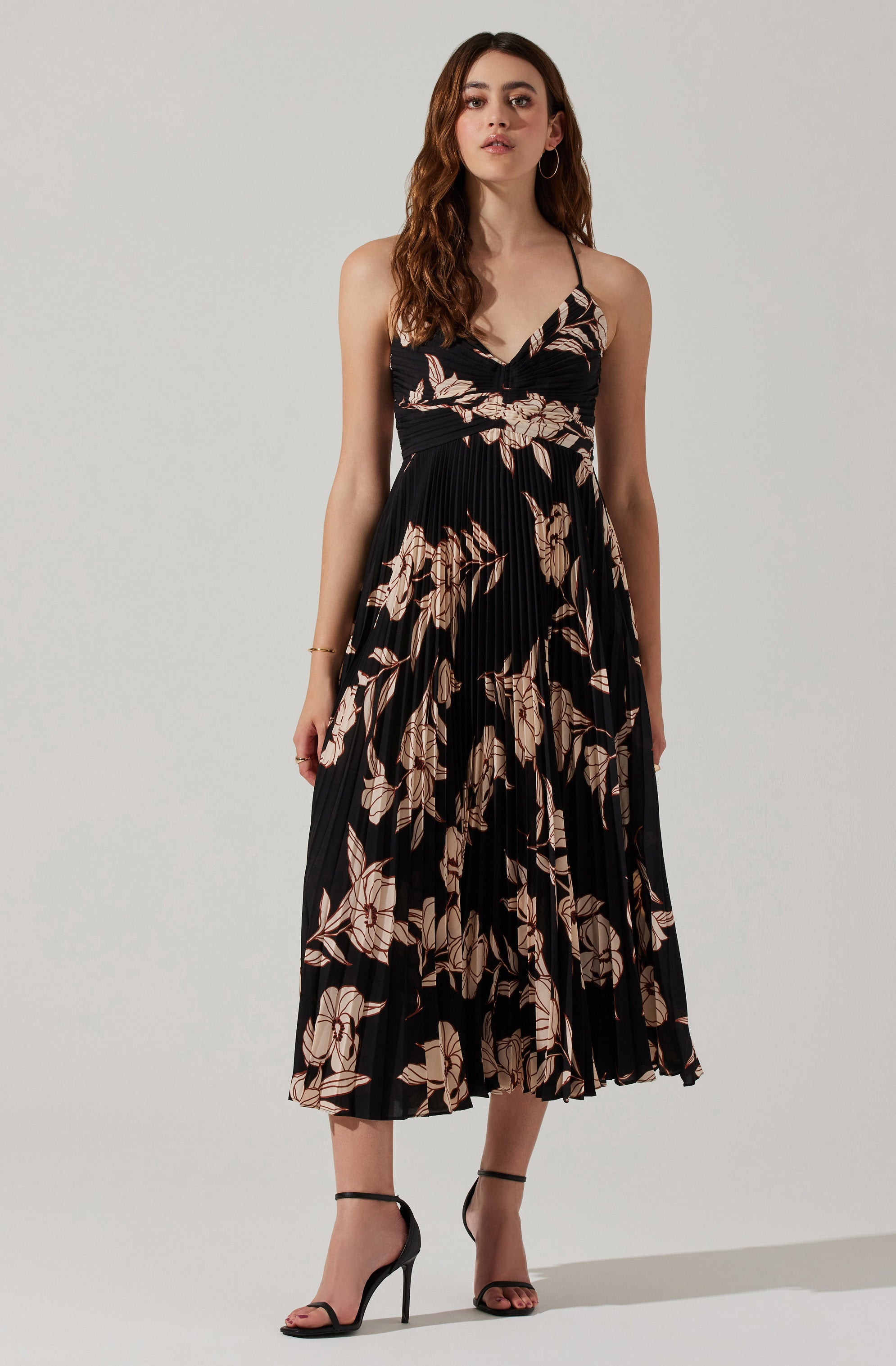 A woman is wearing a sleeveless, black dress with a floral pattern in shades of beige and light brown. The dress has a V-neckline, thin shoulder straps, and a pleated skirt that falls below the knees. She is paired with black high-heeled sandals with ankle straps.