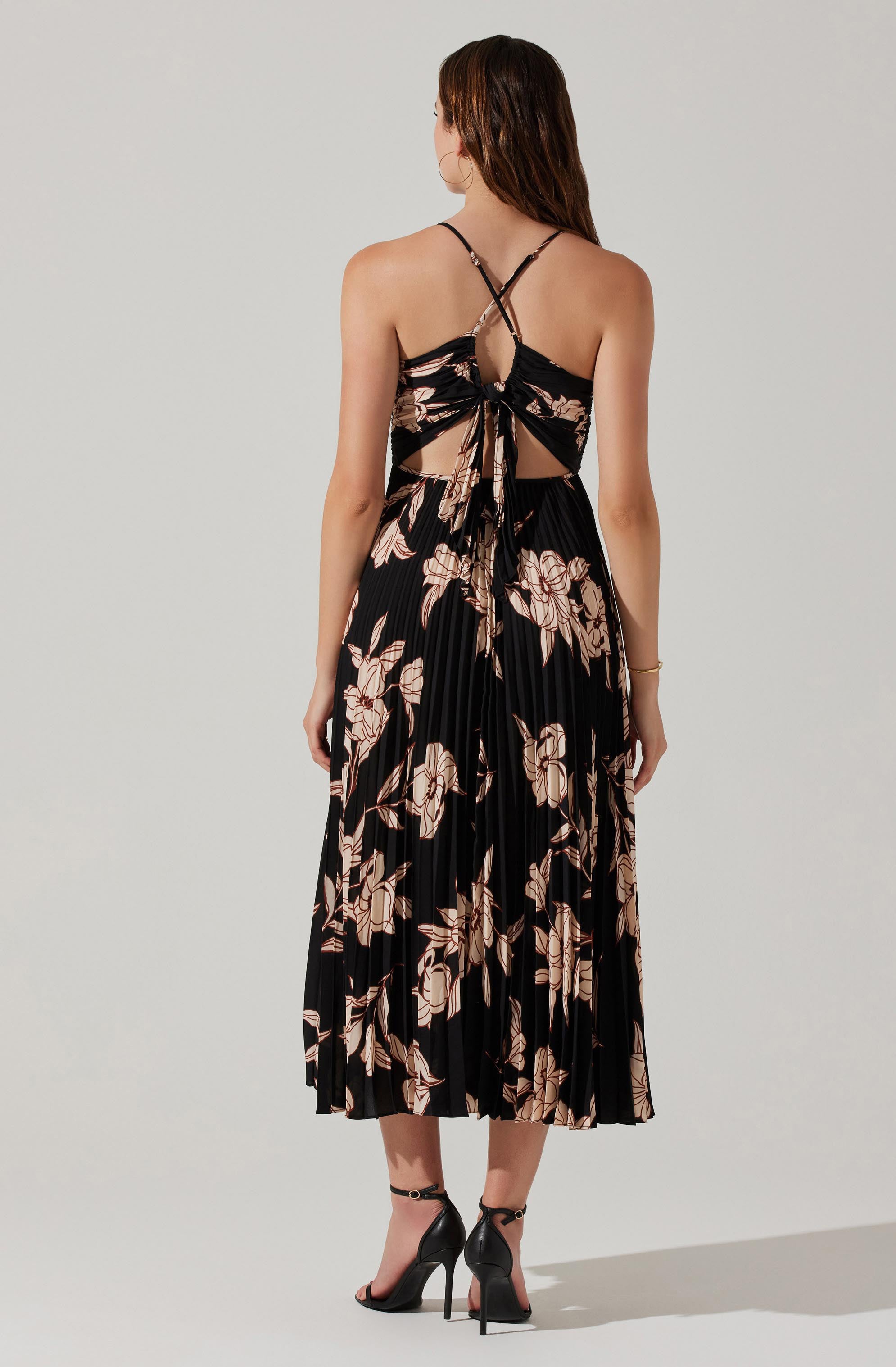 A woman is wearing a black, pleated, floral dress with beige and pinkish floral patterns. The dress features a cut-out back with a tie detail at the waist and thin, crisscross straps over her shoulders. She has on black high-heeled sandals with ankle straps.