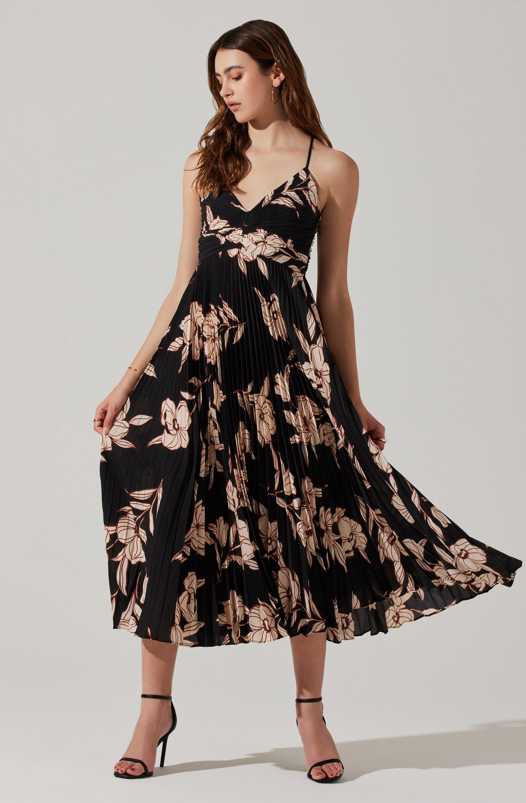 A woman is wearing a sleeveless, black dress with beige and pink floral patterns. The dress has thin spaghetti straps and a fitted bodice, with a pleated skirt that falls below the knees. She is also wearing black high-heeled sandals with ankle straps.