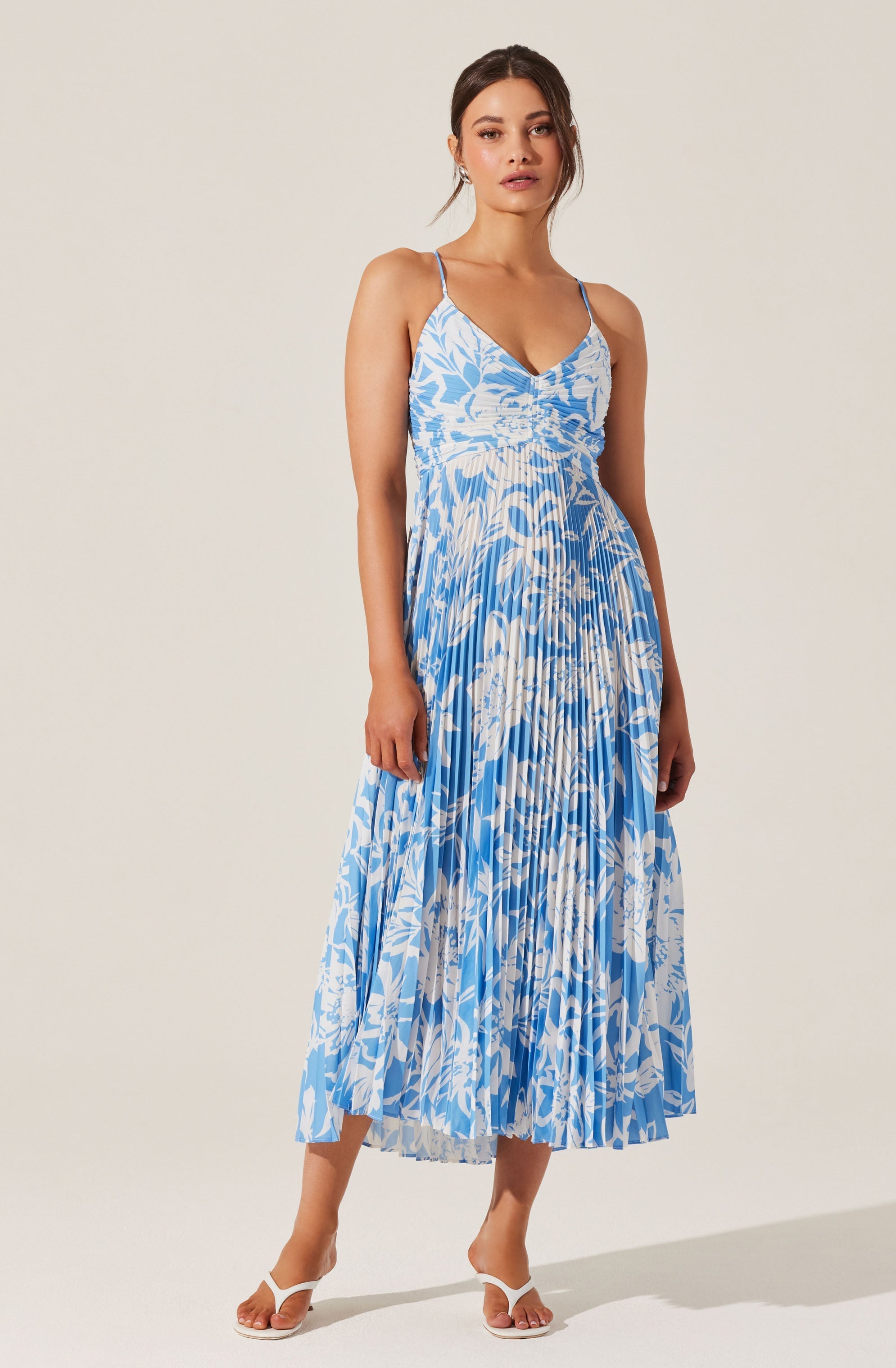 A woman is wearing a sleeveless, flowy maxi dress with thin spaghetti straps. The dress features a blue and white abstract floral pattern with pleats running vertically from top to bottom. She is also wearing white strappy high-heeled sandals.