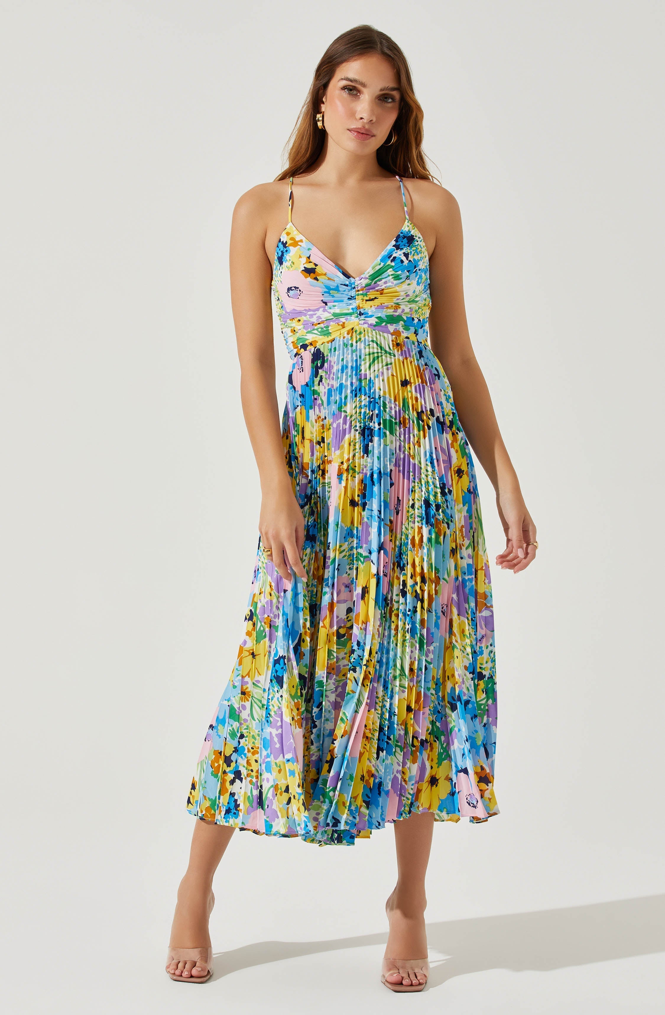 The dress features spaghetti straps and a fitted bodice with a ruched detail at the center. The skirt is long, flowy, and pleated, with a colorful abstract floral pattern in shades of blue, yellow, purple, green, and black.
