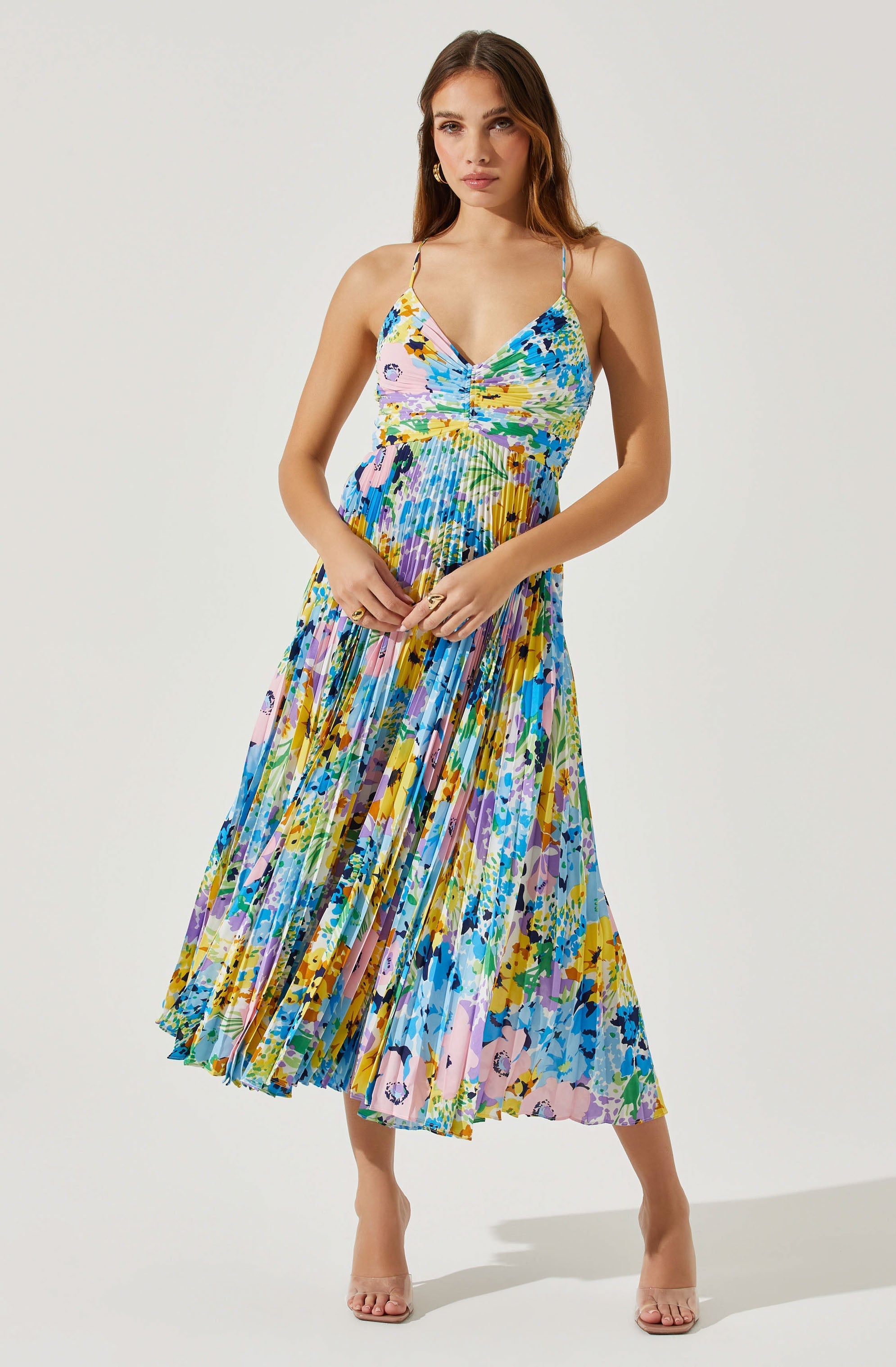 A woman is wearing a sleeveless, ankle-length dress with thin spaghetti straps. The dress features a colorful, abstract floral pattern with shades of blue, pink, yellow, green, purple, and black. The fabric appears lightweight and flowy, with pleats throughout the skirt, creating a vibrant and dynamic look.