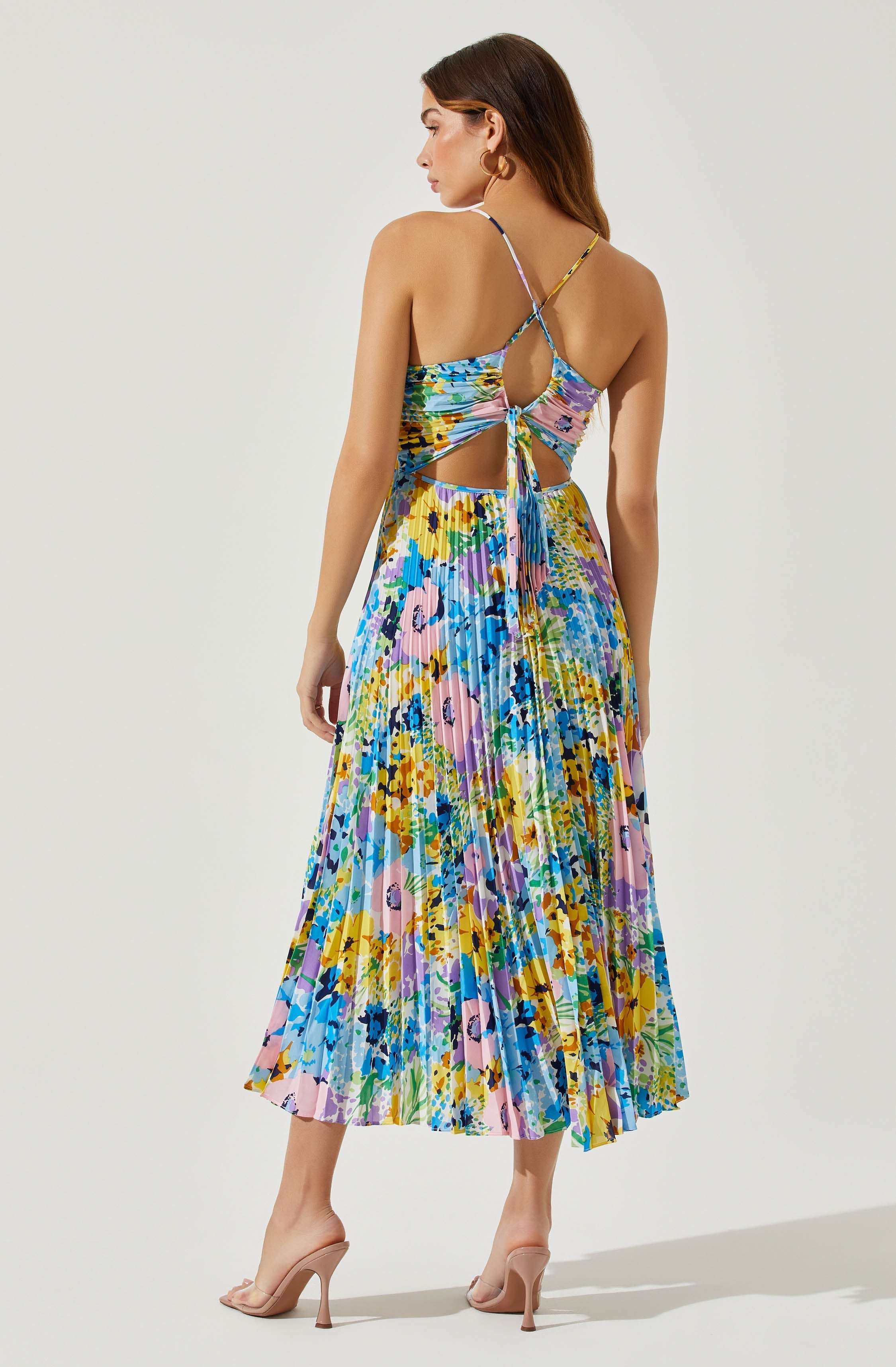 A woman wearing a colorful, floral-patterned dress with a pleated skirt. The dress features thin spaghetti straps, a cutout back with a tie detail, and a high-low hemline. She is also wearing nude high heels.