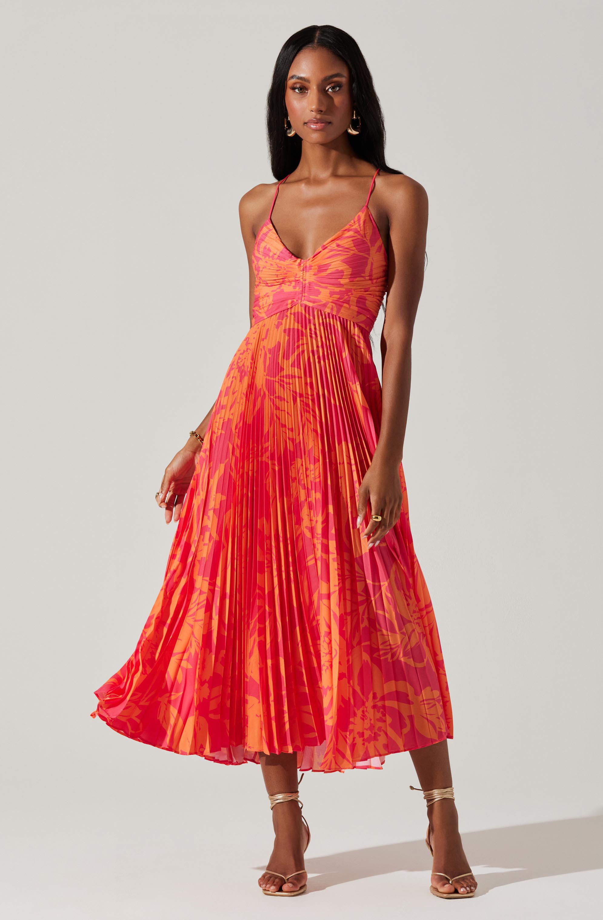 A woman is wearing a sleeveless, midi-length dress with thin spaghetti straps. The dress features a V-neckline and a fitted bodice, with a vibrant, abstract pattern in shades of pink and orange. The skirt has pleats that create a flowing, dynamic appearance. The woman is also wearing strappy high-heeled sandals in a neutral color.