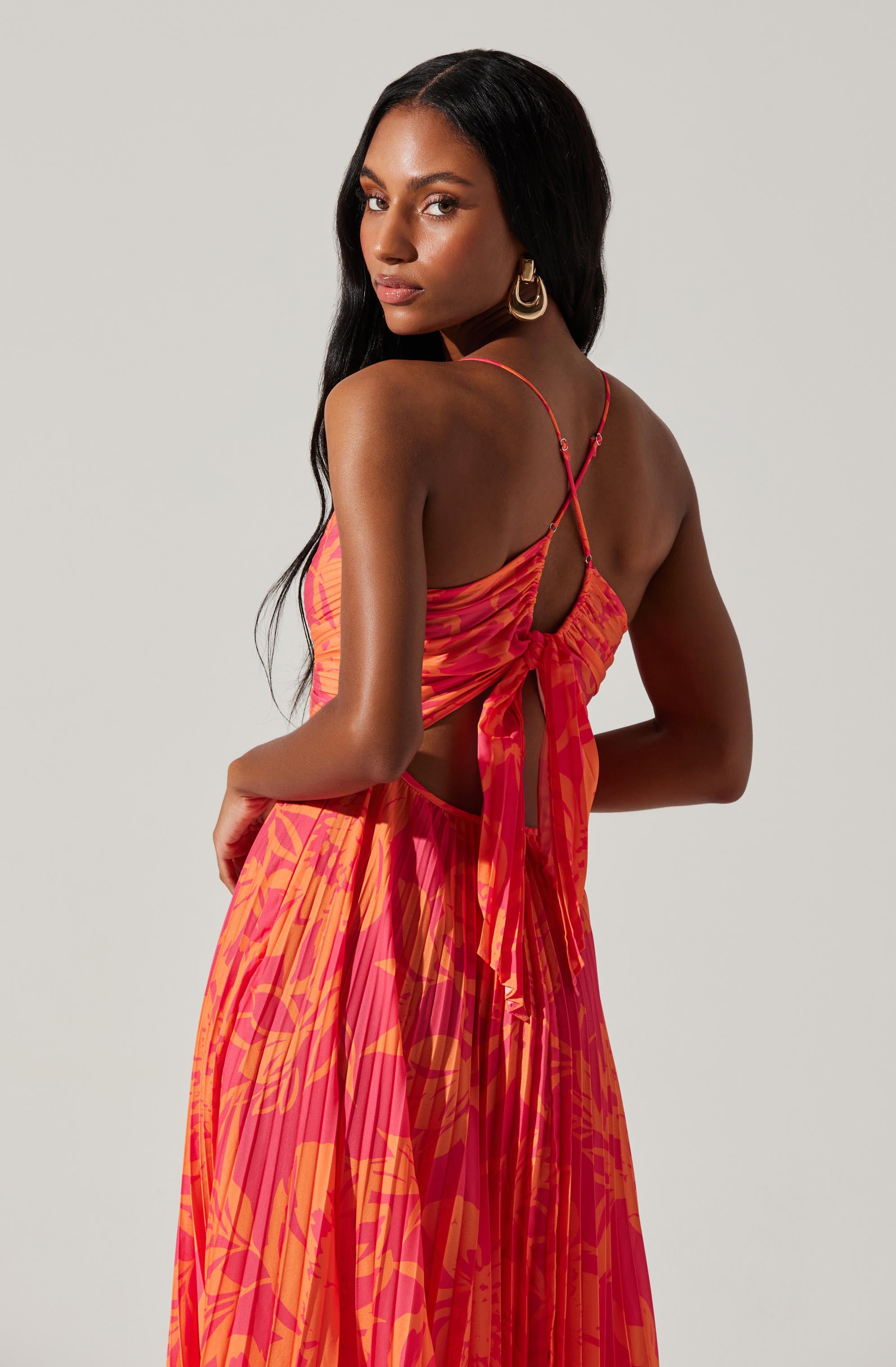 A woman is wearing a vibrant, sleeveless maxi dress with a pink and orange abstract pattern. The dress features thin, adjustable spaghetti straps that crisscross at the back and a large decorative bow tied at the lower back, creating an open-back design. The dress has a flowing, pleated skirt.