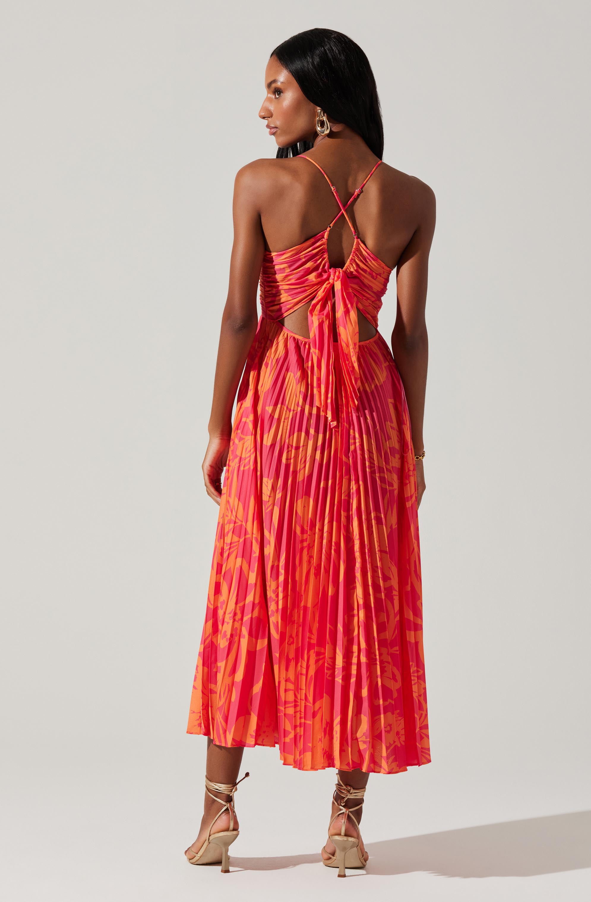 A woman is wearing a vibrant, sleeveless maxi dress with thin spaghetti straps that crisscross at the back. The dress features a ruched bodice tied in a large bow at the mid-back and has cut-outs on the sides. The skirt is pleated and has a bold, abstract pattern in shades of red, orange, and pink. She pairs the dress with beige, strappy high-heeled sandals that wrap around her ankles.