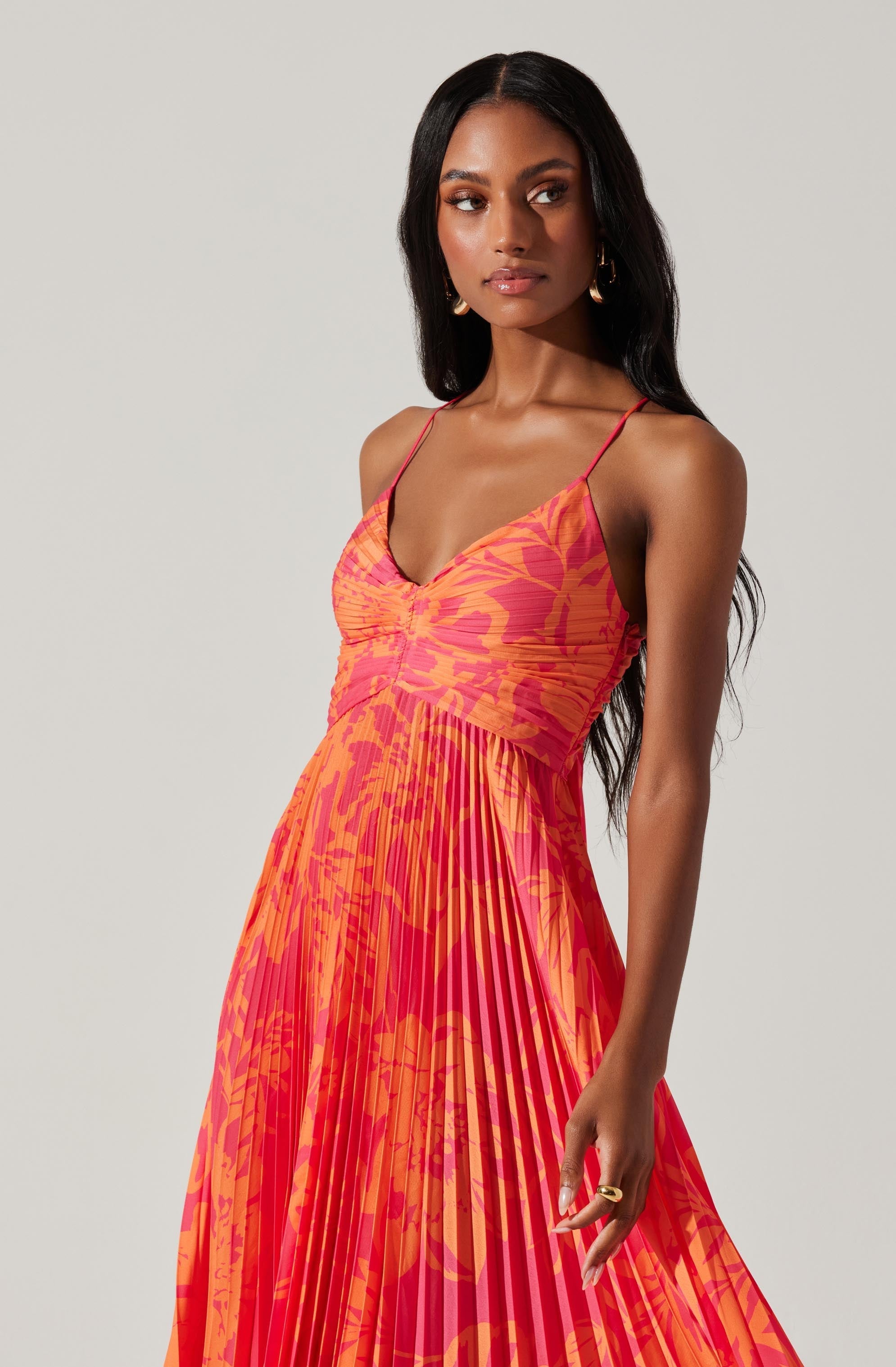 A woman wearing a colorful, sleeveless dress with thin spaghetti straps. The dress features a vibrant orange and pink floral pattern with a ruched bodice and a pleated skirt.