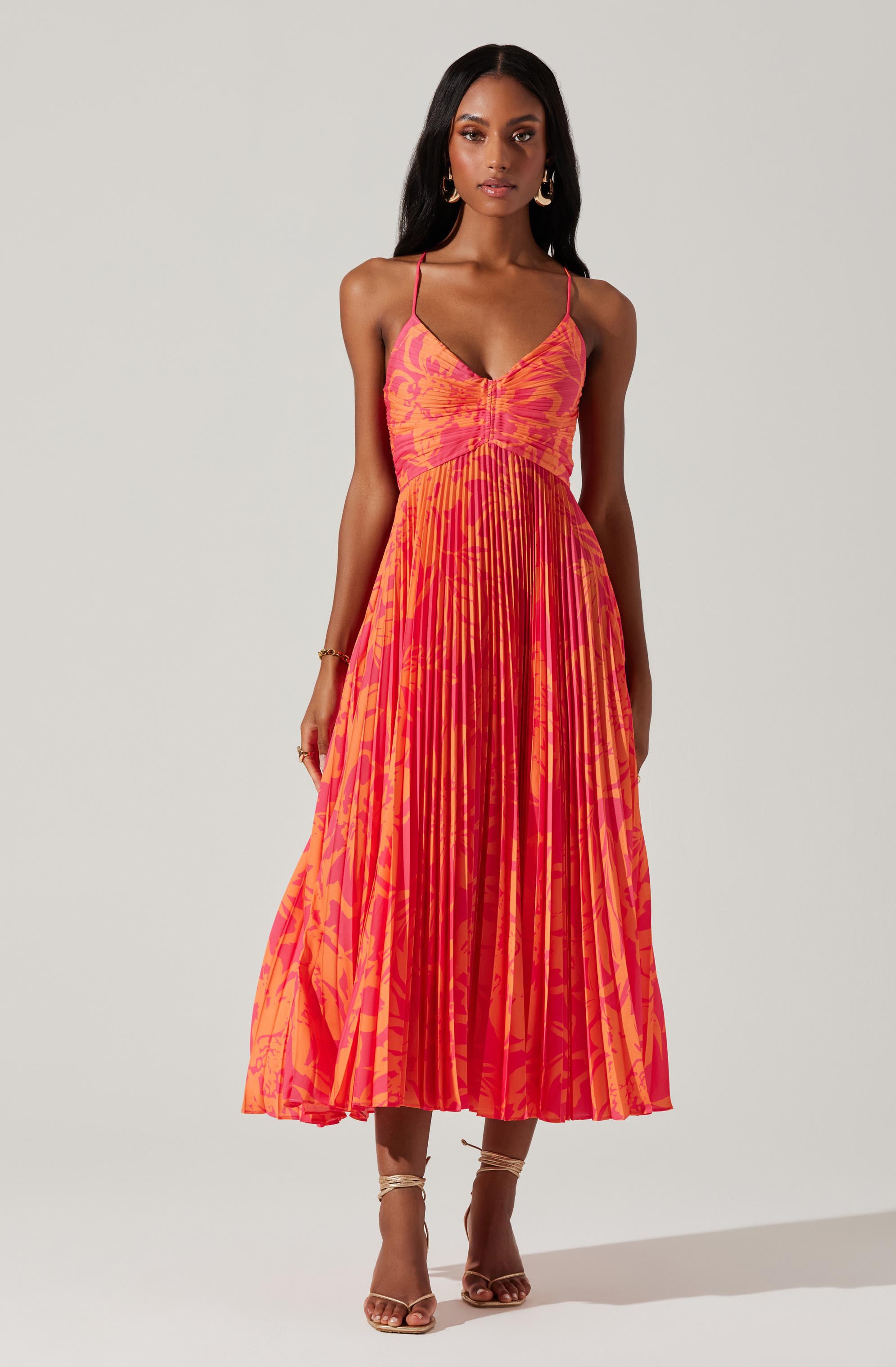 A woman is wearing a long, flowy dress with thin spaghetti straps. The dress features a vibrant pattern of orange and pink hues, with a gathered bodice and pleated skirt. She has on gold hoop earrings, a bracelet, and strappy heels.
