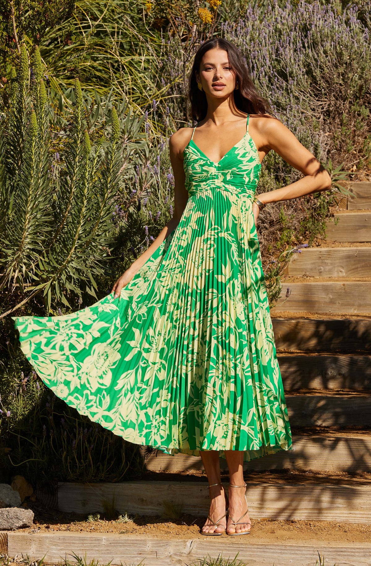 A woman is wearing a sleeveless, ankle-length dress with a green and white tropical floral pattern. The dress features thin shoulder straps, a ruched bodice, and a flowing pleated skirt. She is standing outdoors on wooden steps surrounded by lush greenery and purple flowering plants. She has long, wavy dark hair and is wearing strappy high-heeled sandals.