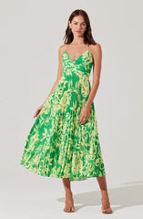A woman is wearing a sleeveless, green and yellow floral dress with a fitted bodice and a flowing, pleated midi skirt. The dress has thin spaghetti straps and a deep V neckline, with the floral pattern featuring large, abstract leaf and flower designs. She pairs the dress with nude high-heeled sandals.
