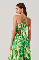 A woman is wearing a sleeveless, green and yellow patterned dress with thin, adjustable spaghetti straps. The dress features a cutout design with a tied knot at the back, creating an open back and sides, and has a flowy, pleated skirt. The fabric has a bold, tropical-inspired leaf pattern. She accessorizes with a large, round earring and a ring on her left hand.