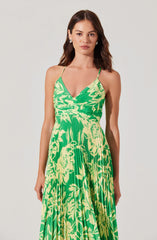A woman is wearing a sleeveless, V-neck maxi dress with thin spaghetti straps. The dress features a vibrant green and yellow floral pattern with pleated fabric.