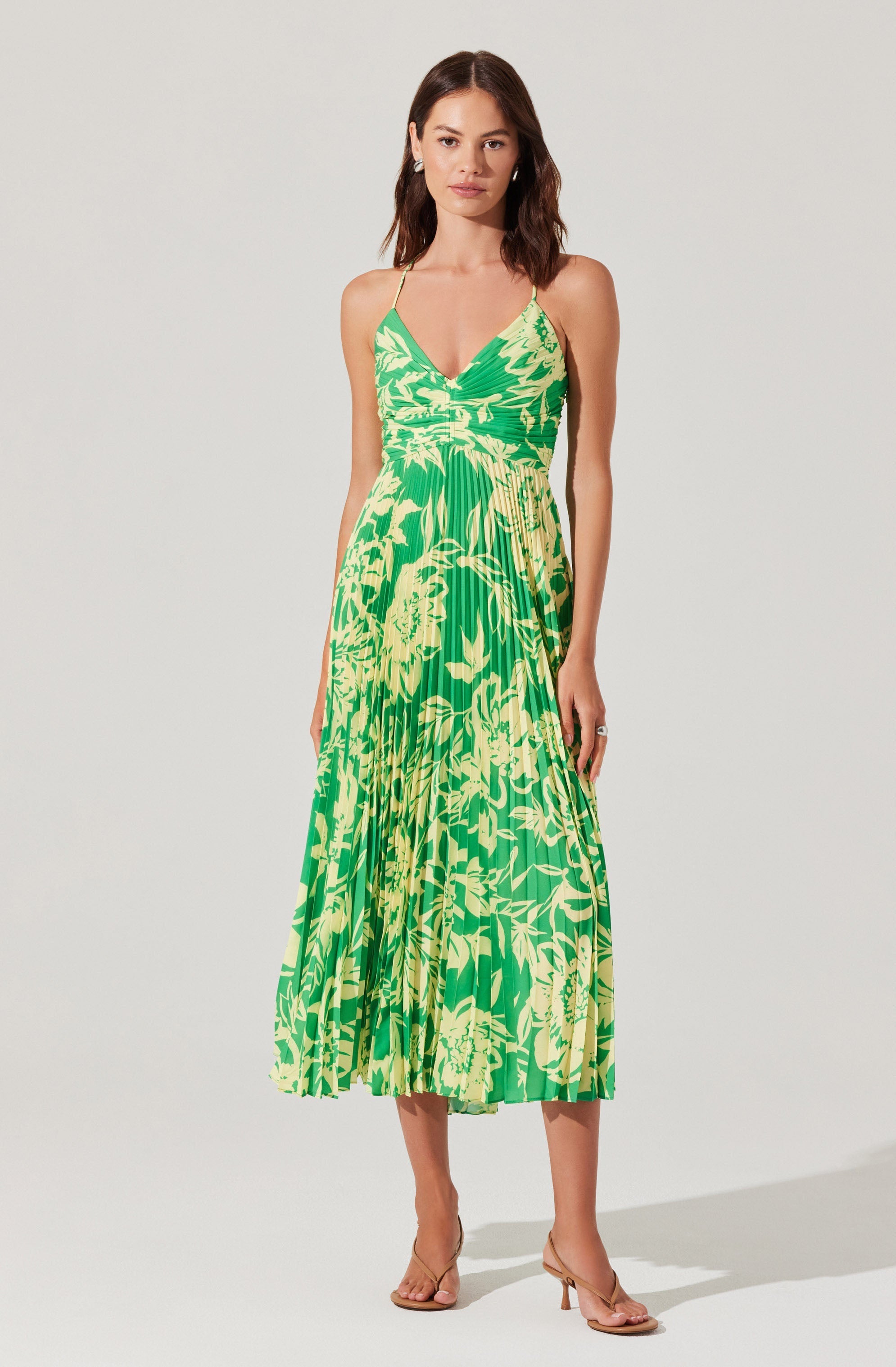 A woman is wearing a sleeveless, V-neck, spaghetti strap dress with a bright green and yellow tropical floral print. The dress has a fitted bodice with ruched detailing at the bust and a pleated, flowy midi-length skirt. She is accessorizing with earrings and a ring, and she is wearing nude high-heeled sandals.