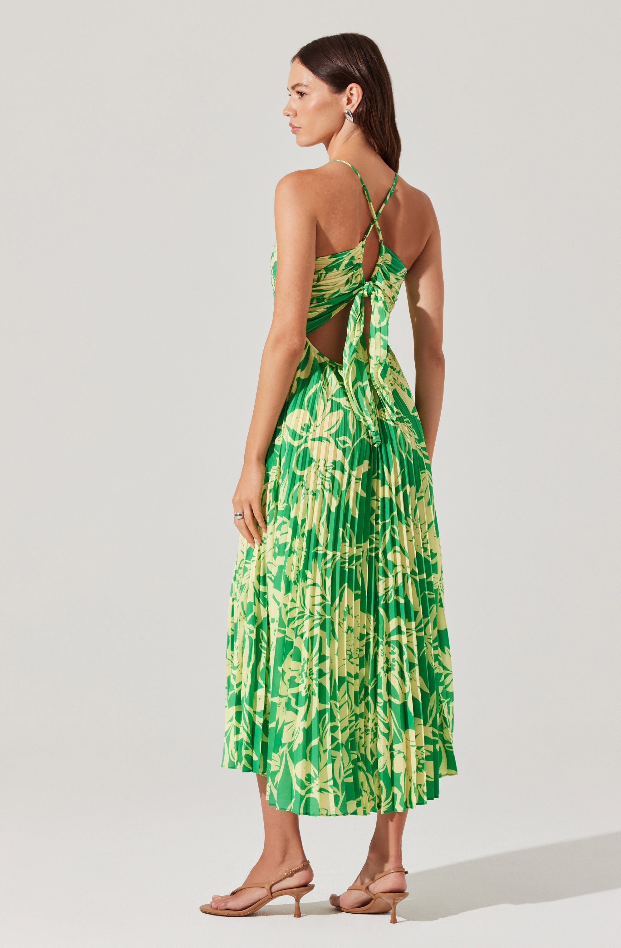 A woman is wearing a vibrant green and yellow floral-printed sleeveless dress with spaghetti straps. The dress features an open back with a tie detail at the lower back and asymmetric cutouts on the sides. She is also wearing nude-colored high-heeled sandals.