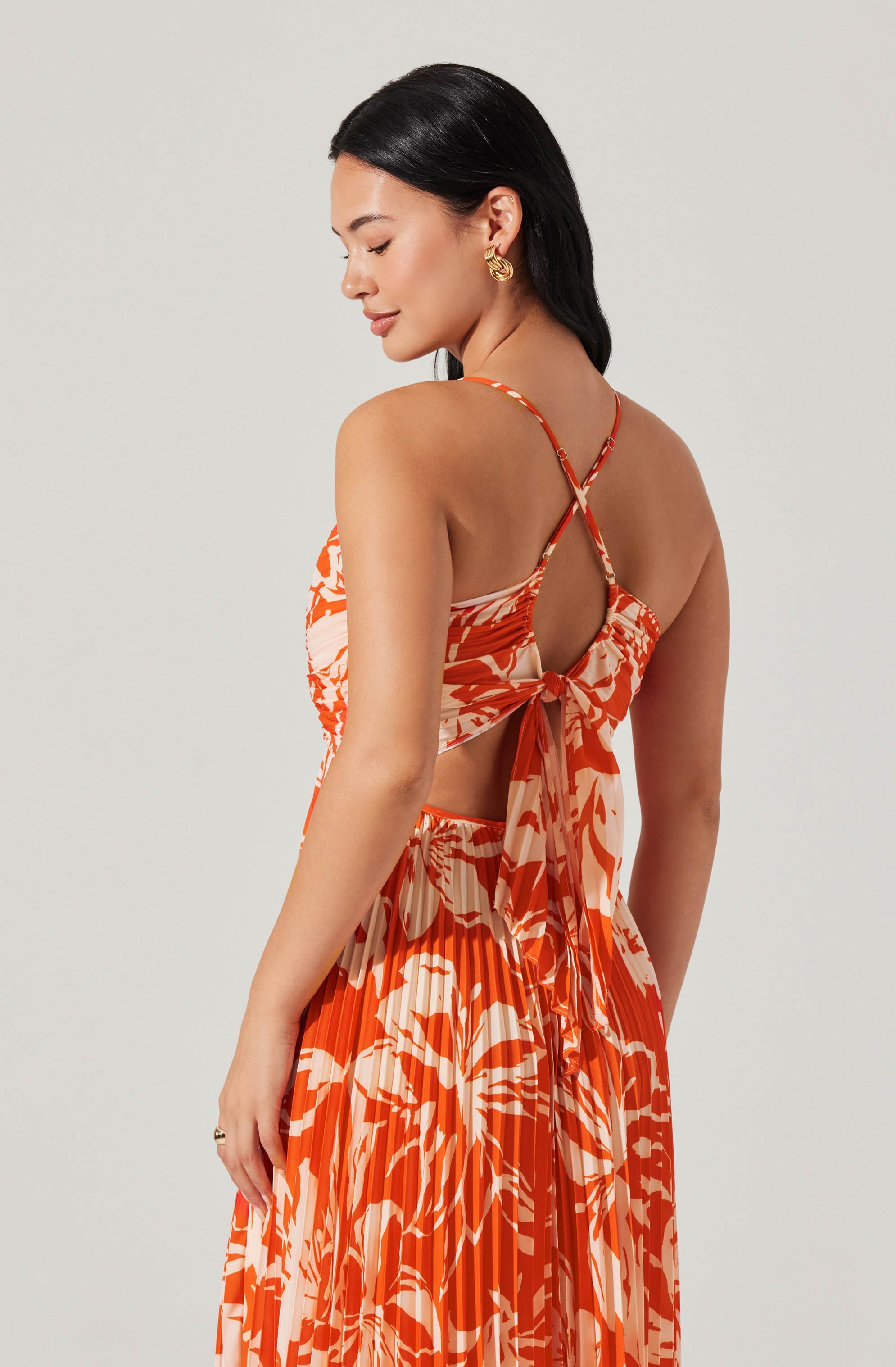 A woman is wearing a matching two-piece outfit with a sleeveless, strappy top and a wide, pleated skirt. The top has thin spaghetti straps and a back tie with a keyhole opening. The fabric features an orange and white abstract, floral print. The pleated skirt is high-waisted and flows down gracefully. She has dark, straight hair and is accessorizing with gold hoop earrings.