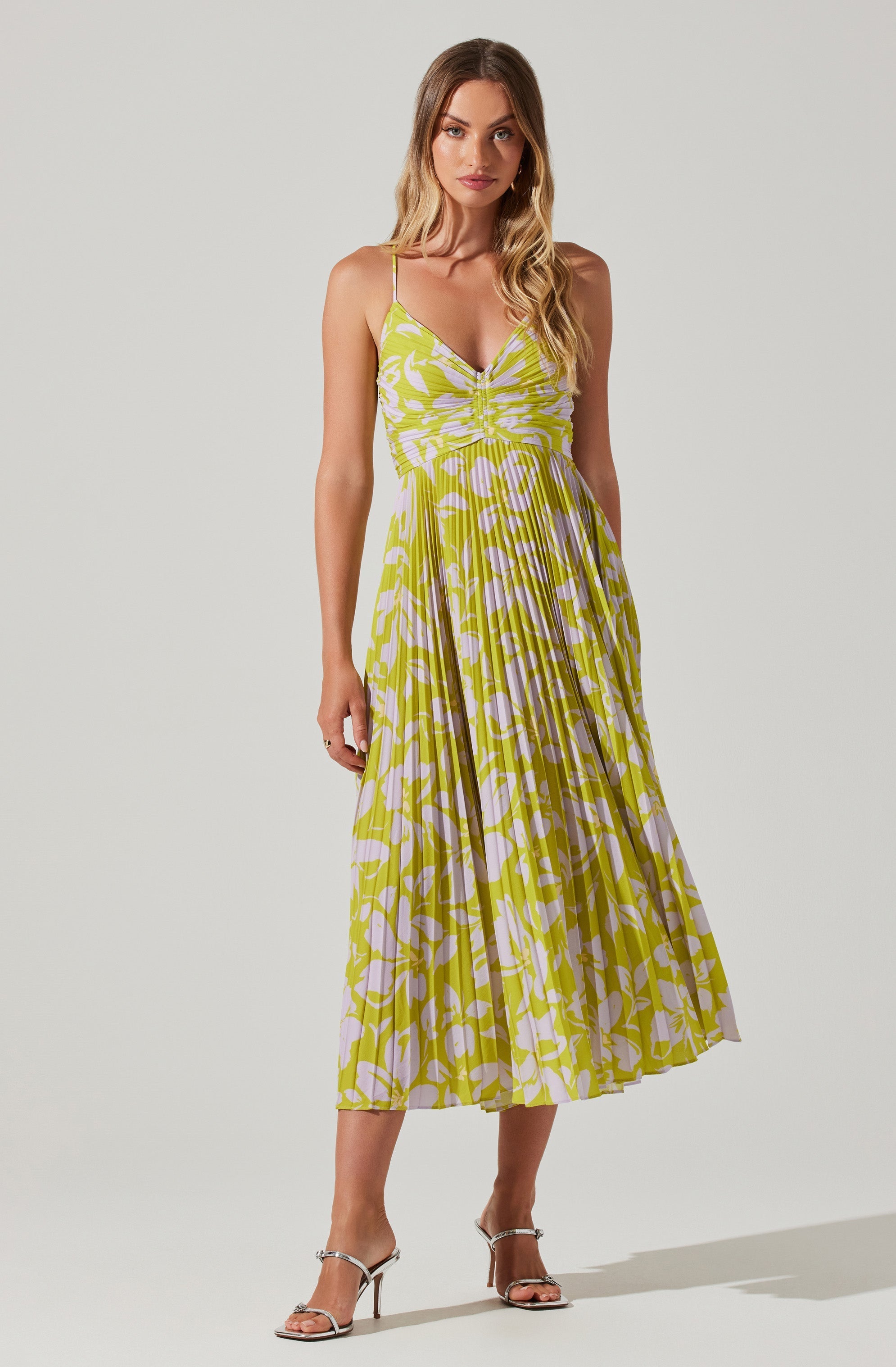 A woman is wearing a sleeveless, knee-length dress with thin spaghetti straps. The dress features a yellow and white abstract floral pattern with a gathered bust and a pleated skirt. She is also wearing high-heeled sandals with thin straps.