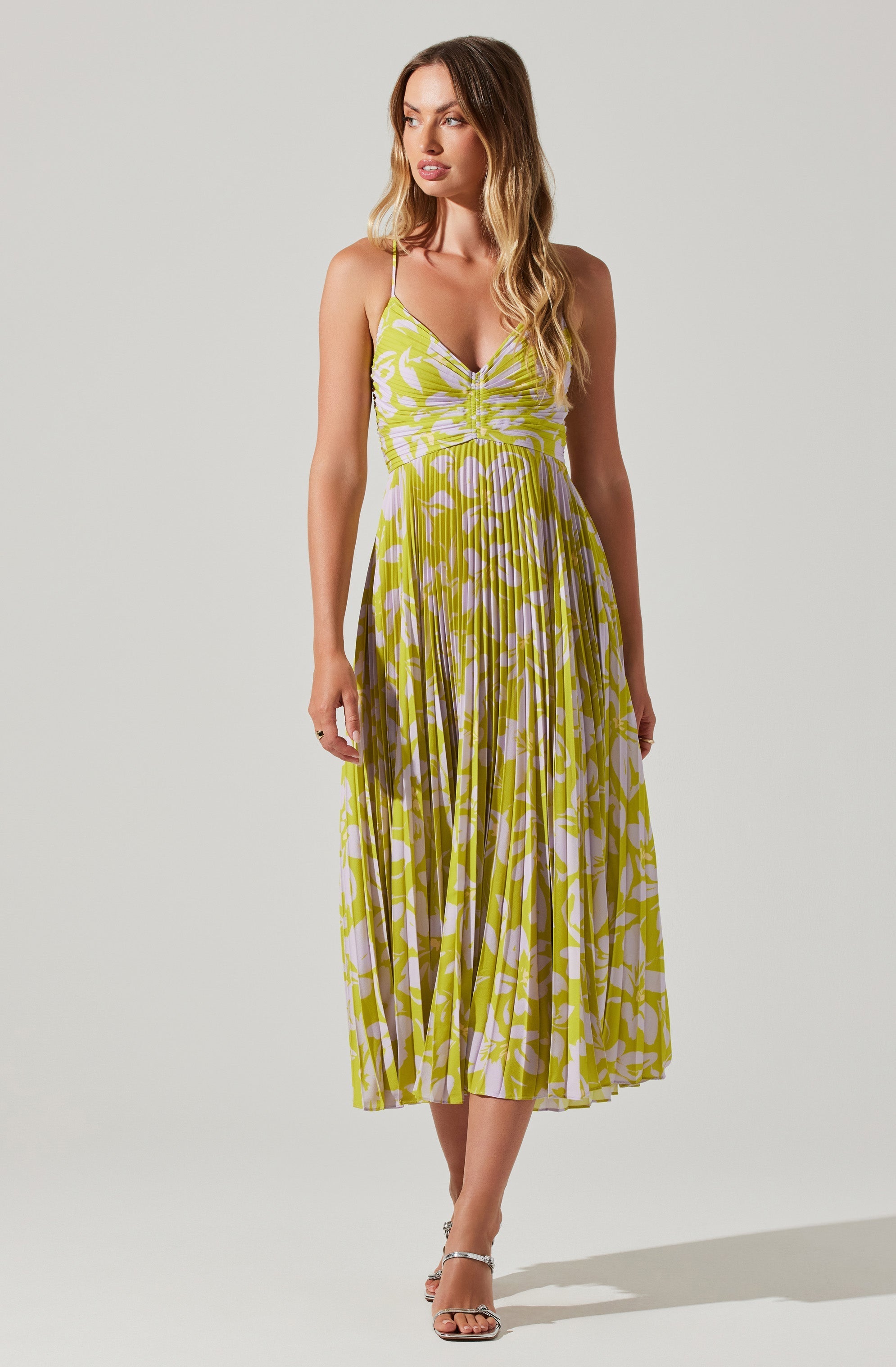 A woman is wearing a sleeveless, V-neck maxi dress with thin spaghetti straps. The dress features a fitted bodice with gathered detail in the center and a flowing skirt with a vibrant, abstract leaf pattern in shades of yellow and white. She pairs the dress with strappy, open-toe high heels.