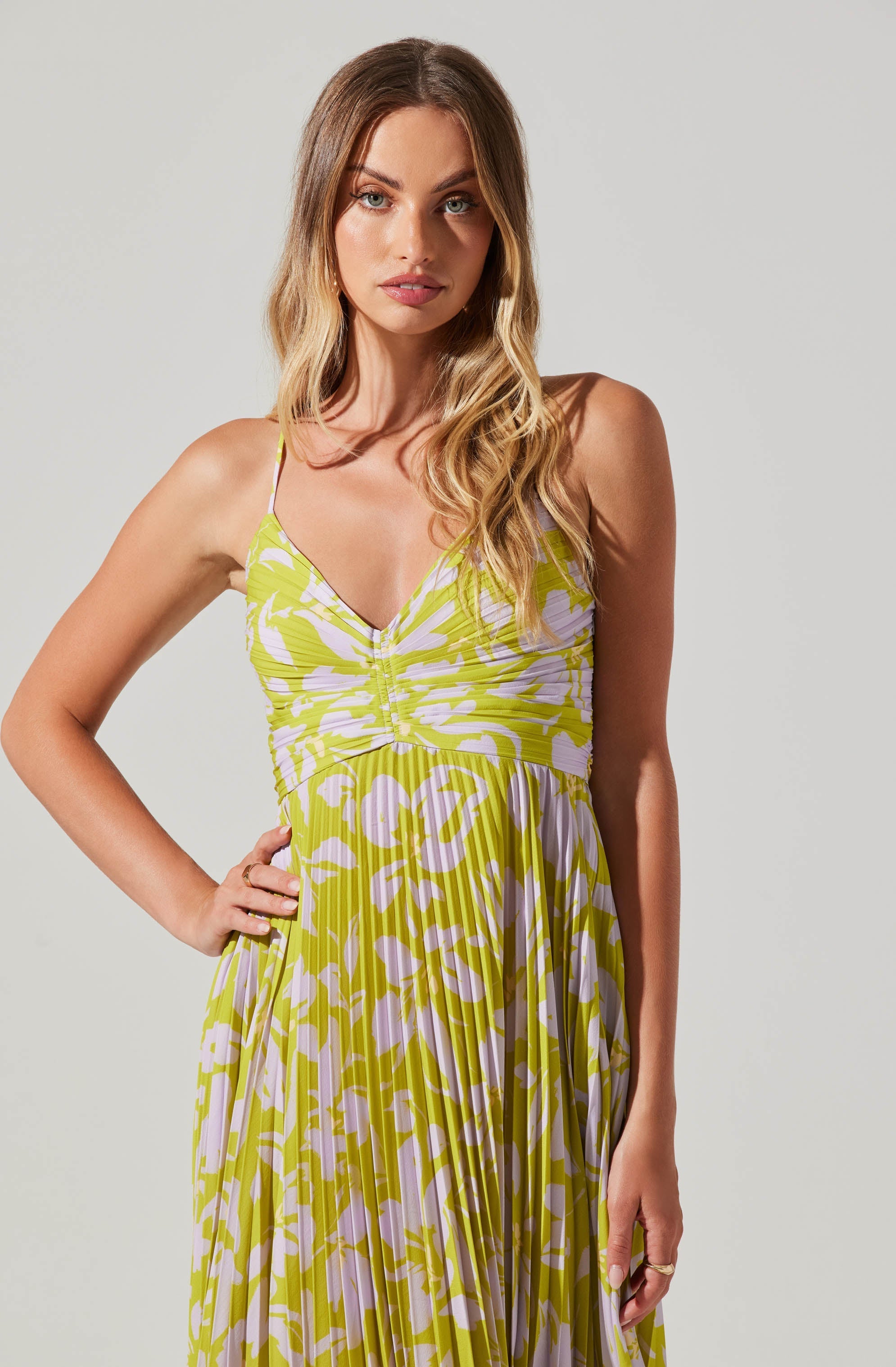 The woman is wearing a sleeveless, V-neck dress with thin spaghetti straps. The dress features a colorful, abstract pattern in shades of yellow and white, with a gathered detail at the bust and a pleated skirt that flows down from the waist.
