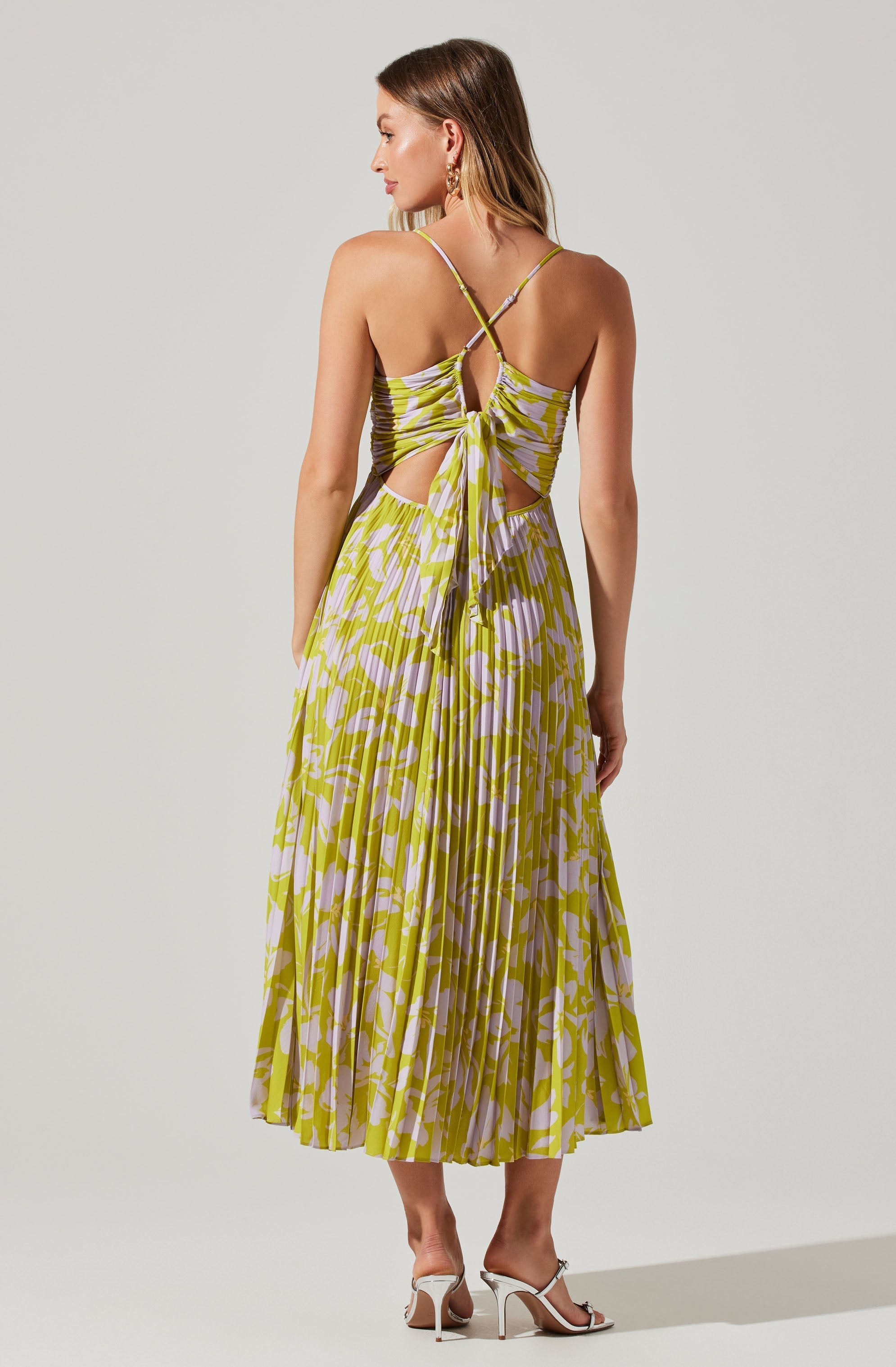 A woman is wearing a sleeveless, flowy dress with a vibrant yellow and white animal print pattern. The dress features thin spaghetti straps, a tie-back detail, and cut-out sections on the sides. She completes her outfit with white high-heeled sandals.