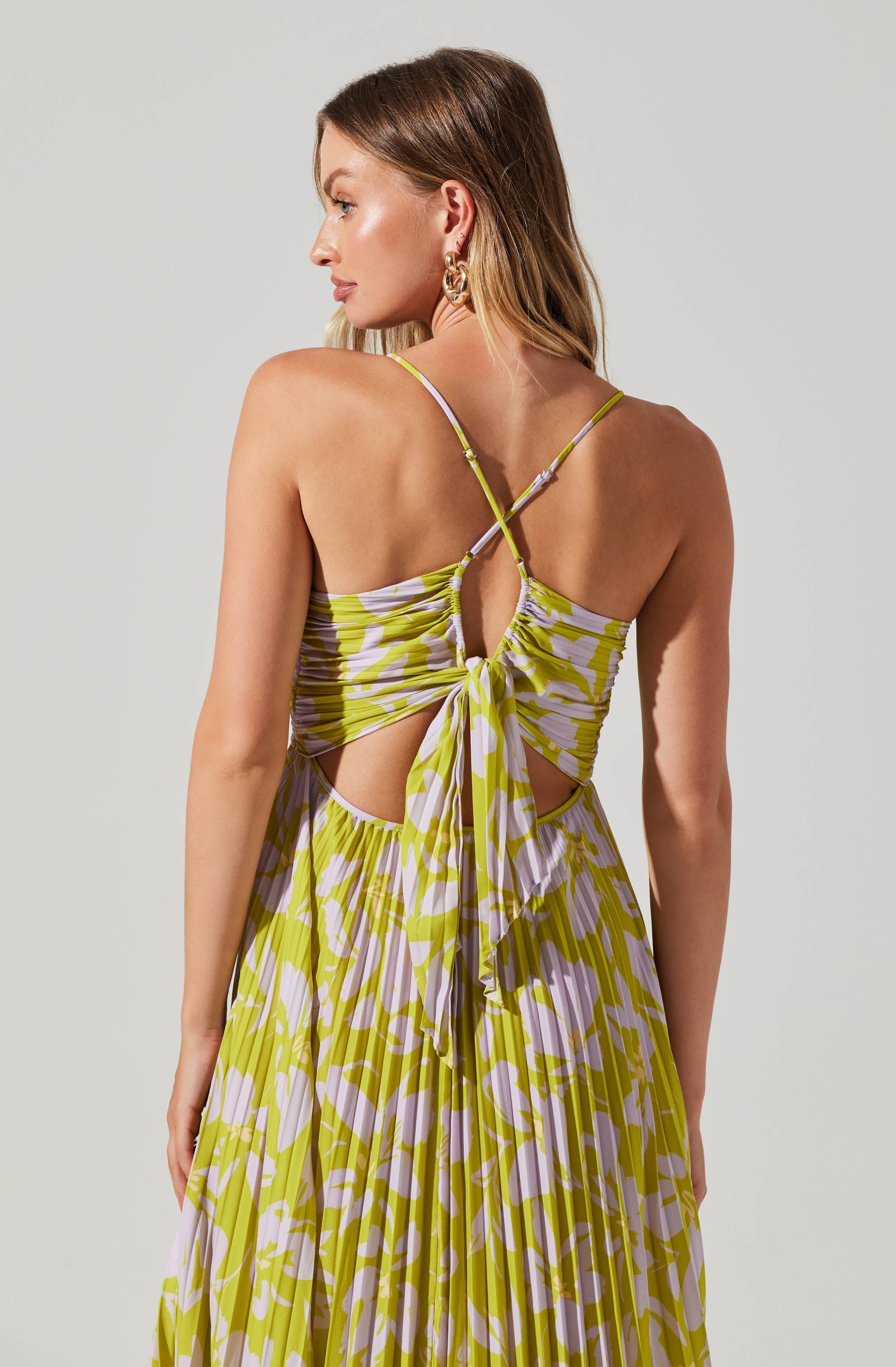 A woman is wearing a sleeveless dress with a yellow and white abstract pattern. The dress features thin, adjustable spaghetti straps that crisscross at the back and an open back with a large bow tied at the waist. The fabric appears lightweight and flowy, suitable for warm weather.