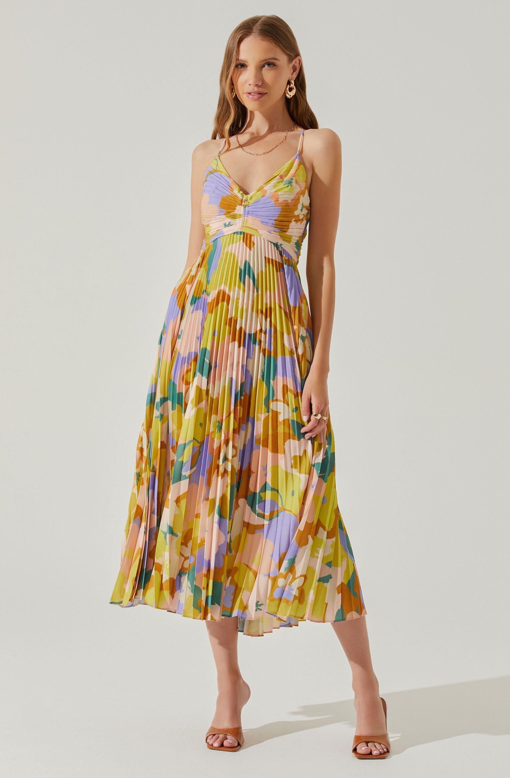 A woman is wearing a sleeveless, V-neck, spaghetti strap dress with a colorful, abstract floral pattern. The dress features a fitted bodice with a ruched detail at the center and a flowing, pleated skirt that falls to mid-calf length. She accessorizes with gold hoop earrings, a layered gold necklace, and rings, and she is wearing tan open-toe heels.