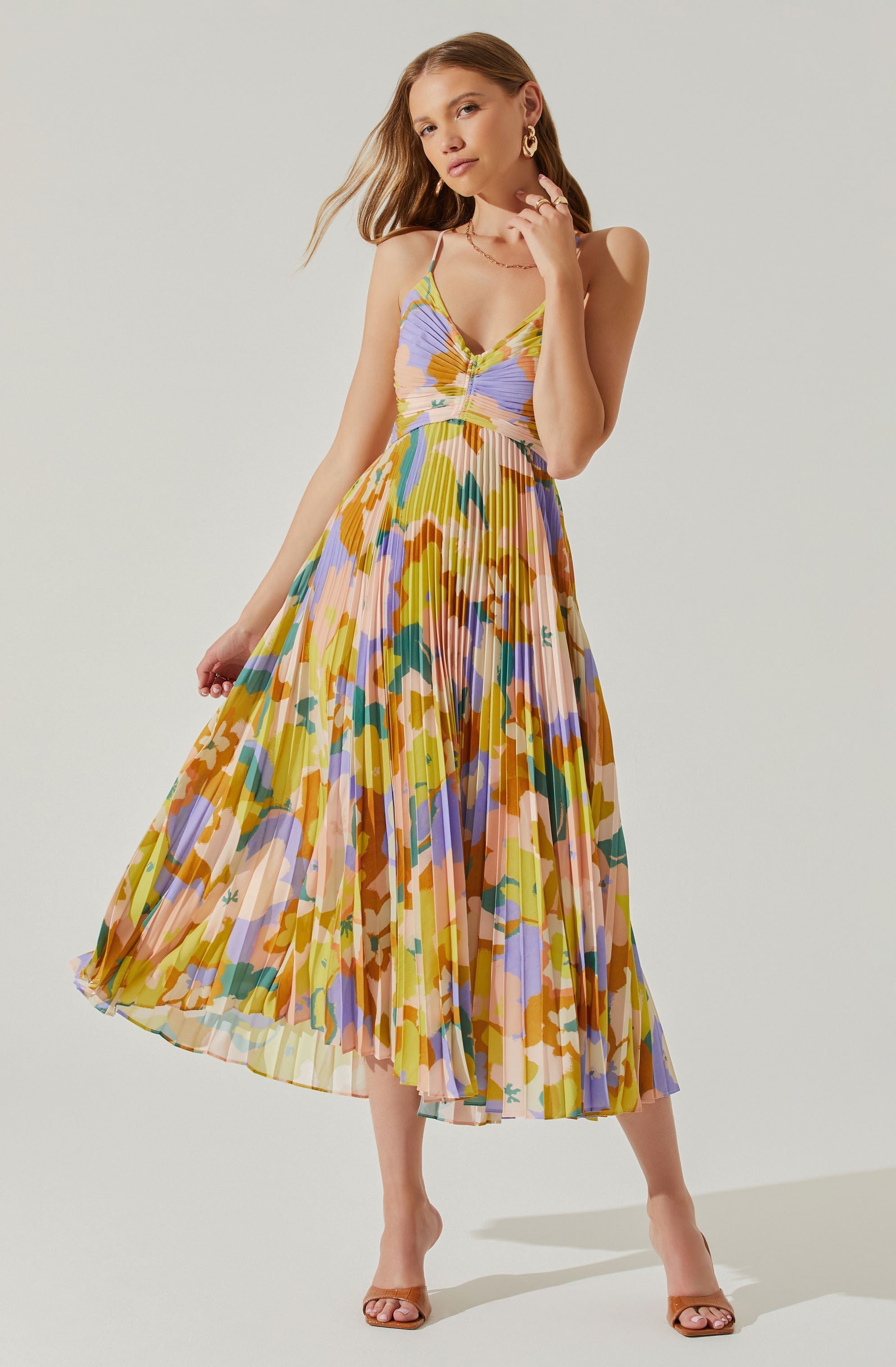 A woman is wearing a flowing, pleated dress with a vibrant, multicolored abstract floral pattern. The dress features thin spaghetti straps and a fitted bodice that gathers at the center.