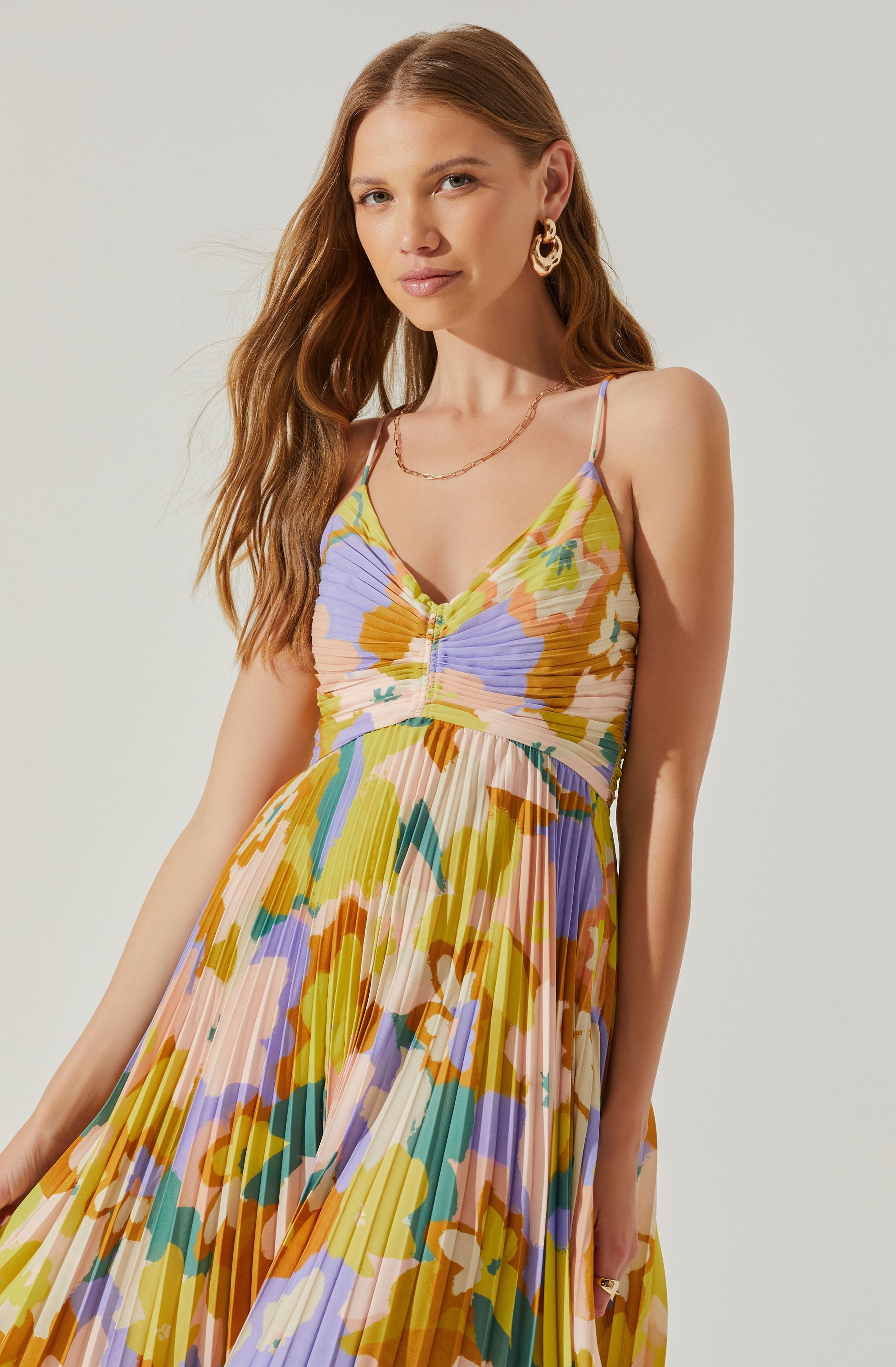 A woman is wearing a sleeveless, V-neck maxi dress with thin spaghetti straps. The dress features a colorful, floral abstract pattern with shades of pastel yellow, purple, green, orange, and cream. The fabric is pleated, creating a textured, flowing appearance.