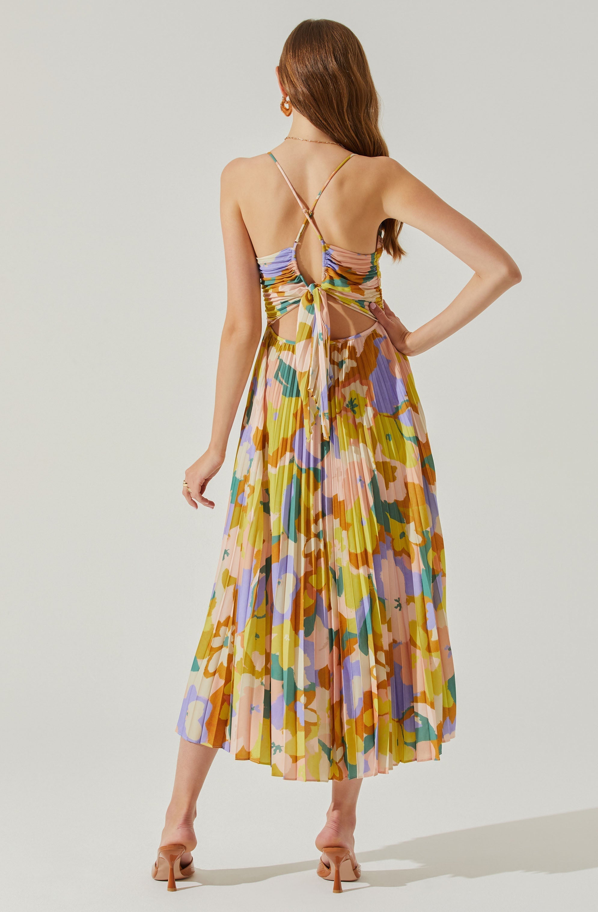 A woman is wearing a colorful, floral-patterned dress with a pleated midi skirt. The dress features a backless design with thin spaghetti straps that cross over her shoulders and tie at the lower back, exposing her back and sides. The fabric has a vibrant mix of pastel and bright colors including yellow, pink, purple, green, and orange. She is also wearing beige high-heeled sandals.