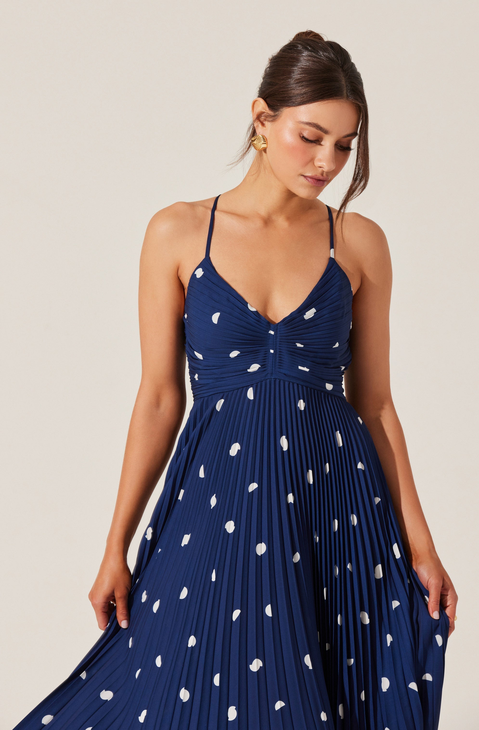 A woman is wearing a dark blue, pleated dress with white polka dots. The dress has thin spaghetti straps and a fitted, ruched bodice. The skirt is long and flowing, with a high waistline. She is accessorized with large, gold statement earrings. Her hair is styled in an elegant updo with loose strands framing her face.