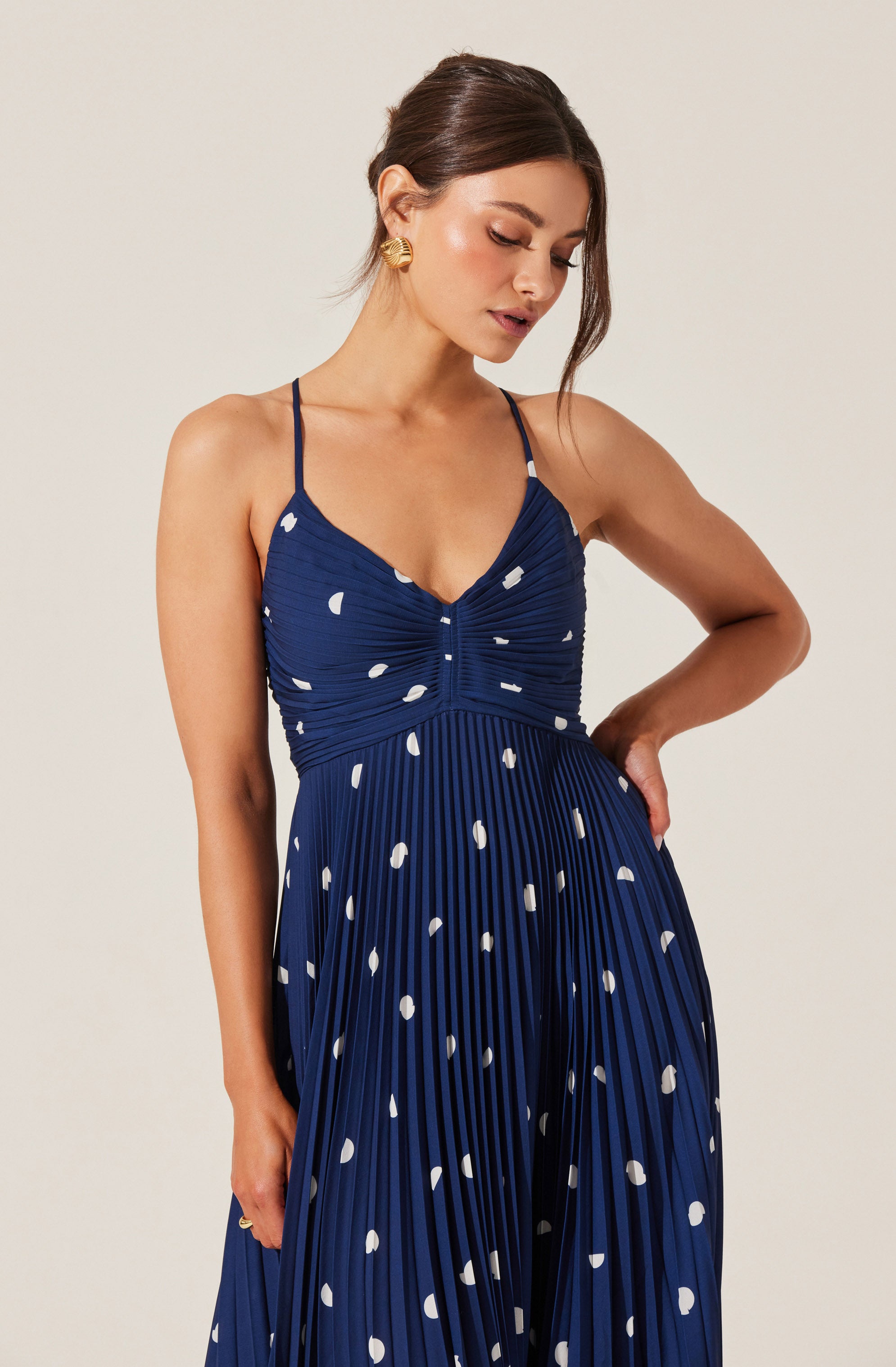 A woman is wearing a blue dress with thin spaghetti straps. The dress features a ruched bodice and a pleated, wide-legged skirt, both decorated with white polka dots.