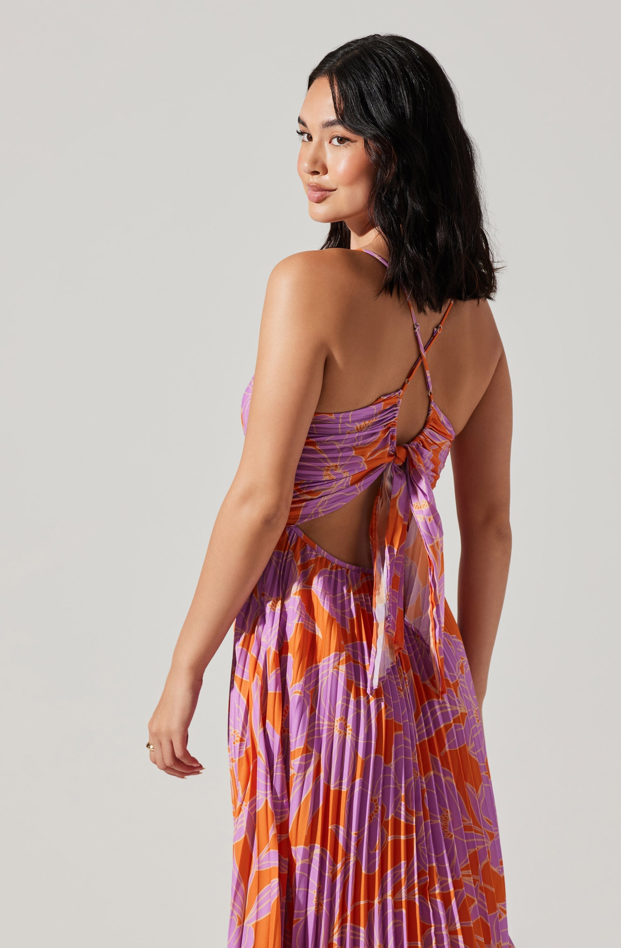 A woman is wearing a flowing, sleeveless maxi dress with a vibrant, abstract floral pattern in shades of purple, orange, and pink. The dress features thin straps crisscrossing at the back and a tie detail at the waist, with a cutout on the side revealing skin.
