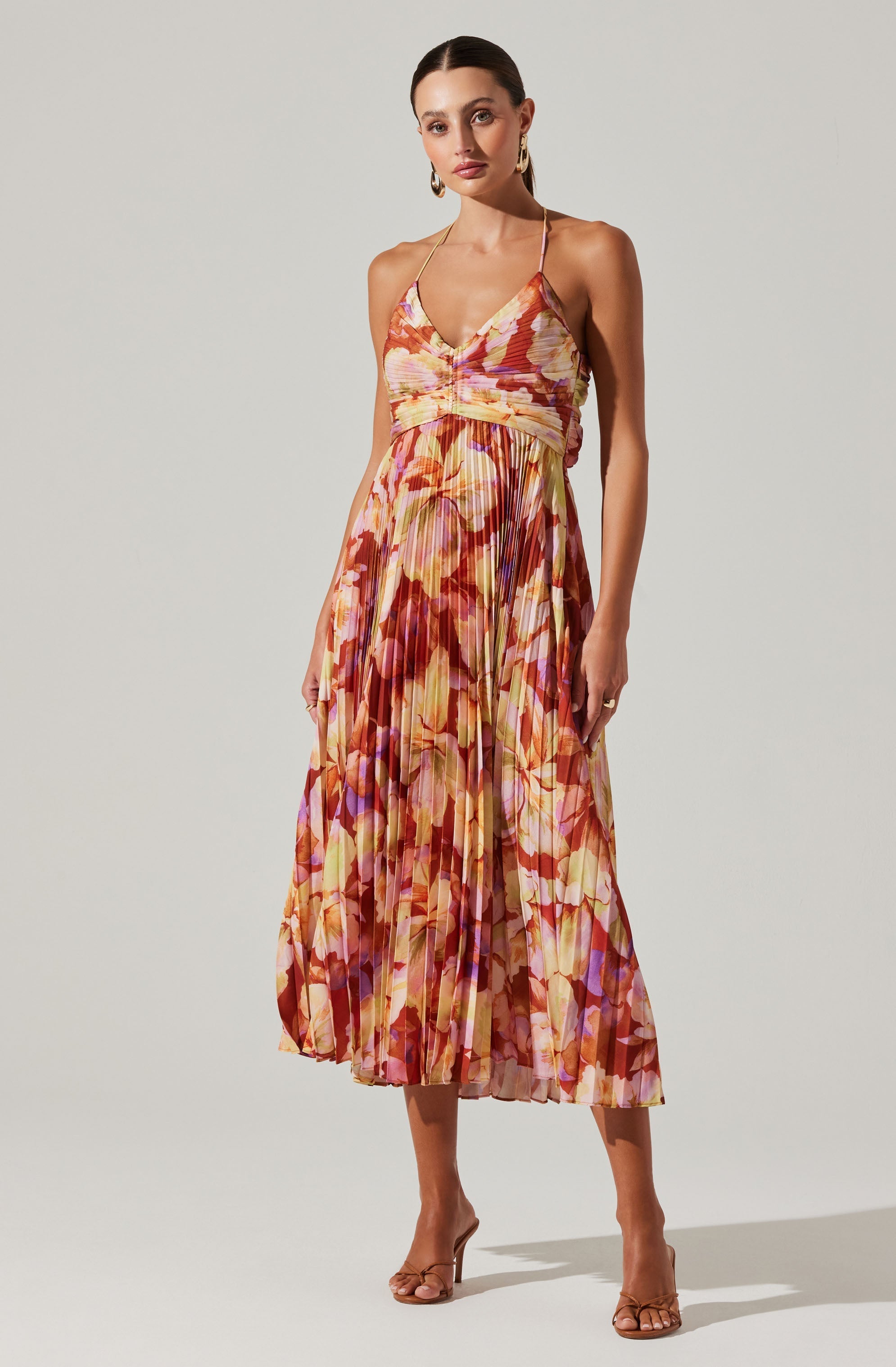 A woman is wearing a sleeveless, floral-printed dress with a fitted bust, spaghetti straps, and a pleated skirt. The dress features warm tones including pink, orange, yellow, and purple, creating a vibrant, colorful pattern. She is also wearing open-toe high heels with thin straps and gold hoop earrings.