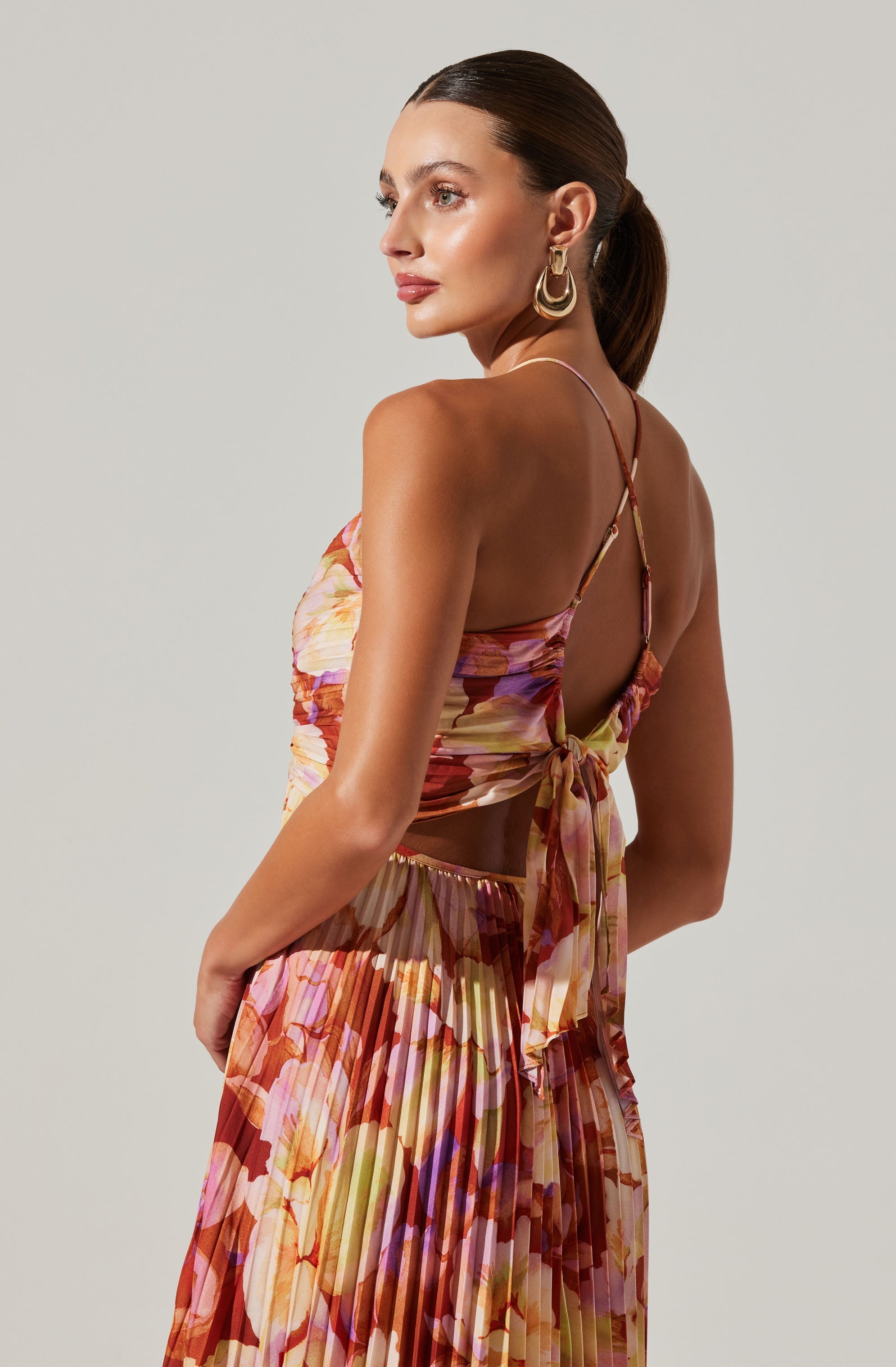 A woman is wearing a floral, sleeveless, pleated dress with a tie at the back. The dress features a mix of warm colors including shades of pink, orange, and yellow. She is accessorized with large, gold hoop earrings.