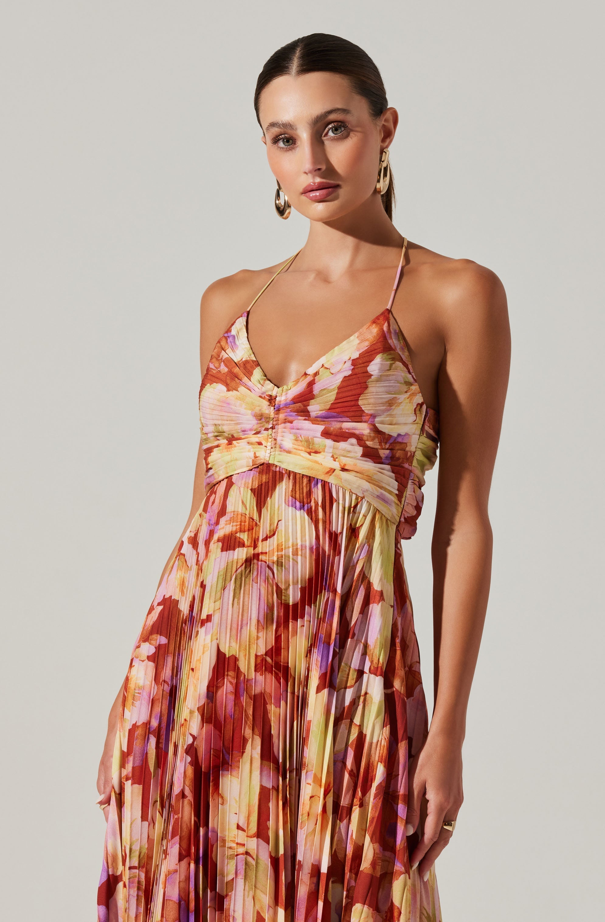 A woman is wearing a sleeveless dress with thin spaghetti straps. The dress features a vibrant floral pattern with shades of orange, pink, purple, yellow, and green, and has a pleated design. The bodice is fitted with a ruched detail in the center, and the skirt flows freely. She has styled her hair in a sleek, pulled-back look and is accessorized with large gold earrings and a gold ring on her right hand.