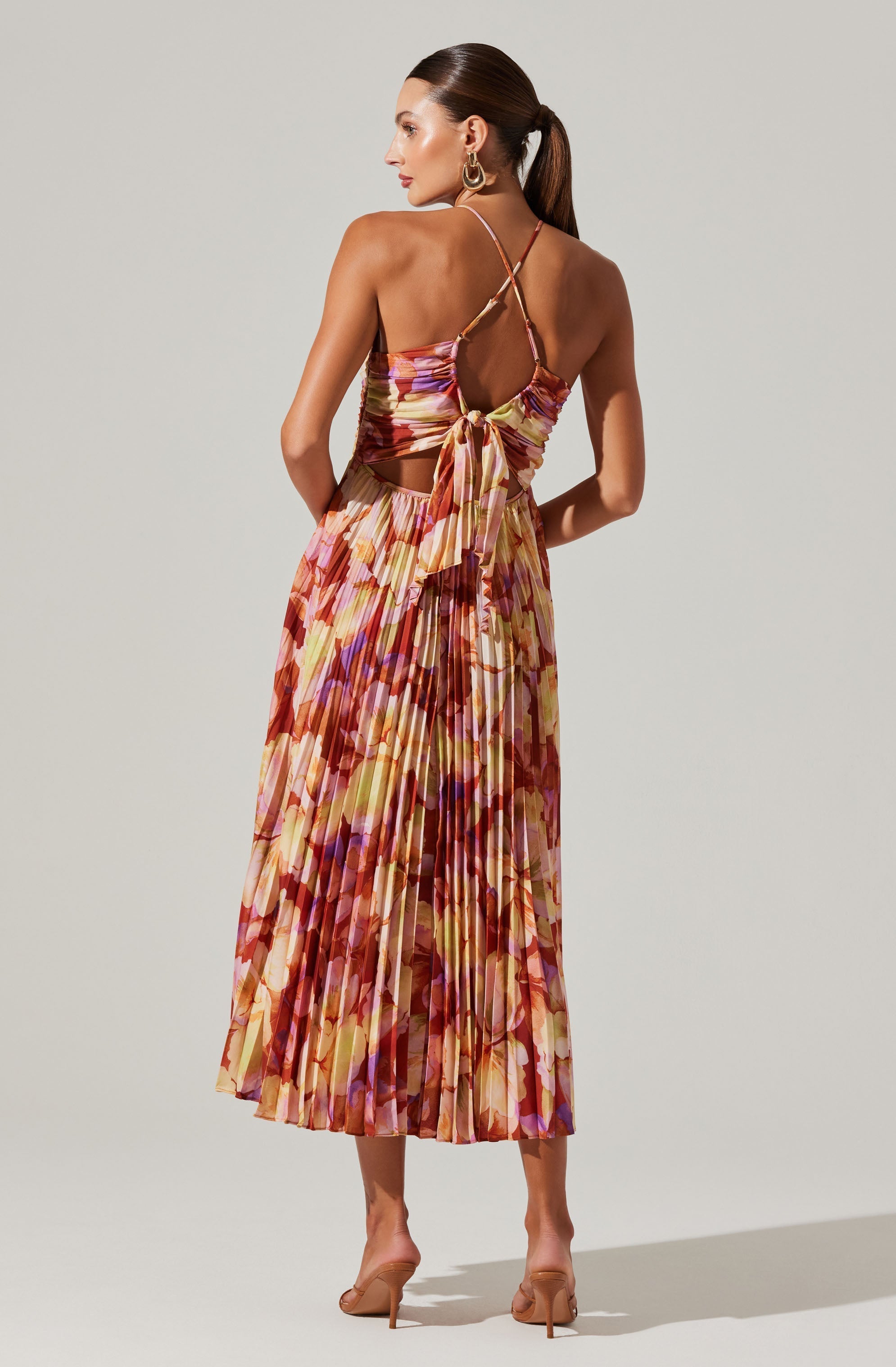 A woman is wearing a colorful pleated dress with a floral pattern in shades of orange, pink, purple, and yellow. The dress features a fitted top with thin spaghetti straps crossing at the back and a large bow tied at the waist, revealing an open back. She is accessorized with large gold hoop earrings and nude high-heeled shoes.