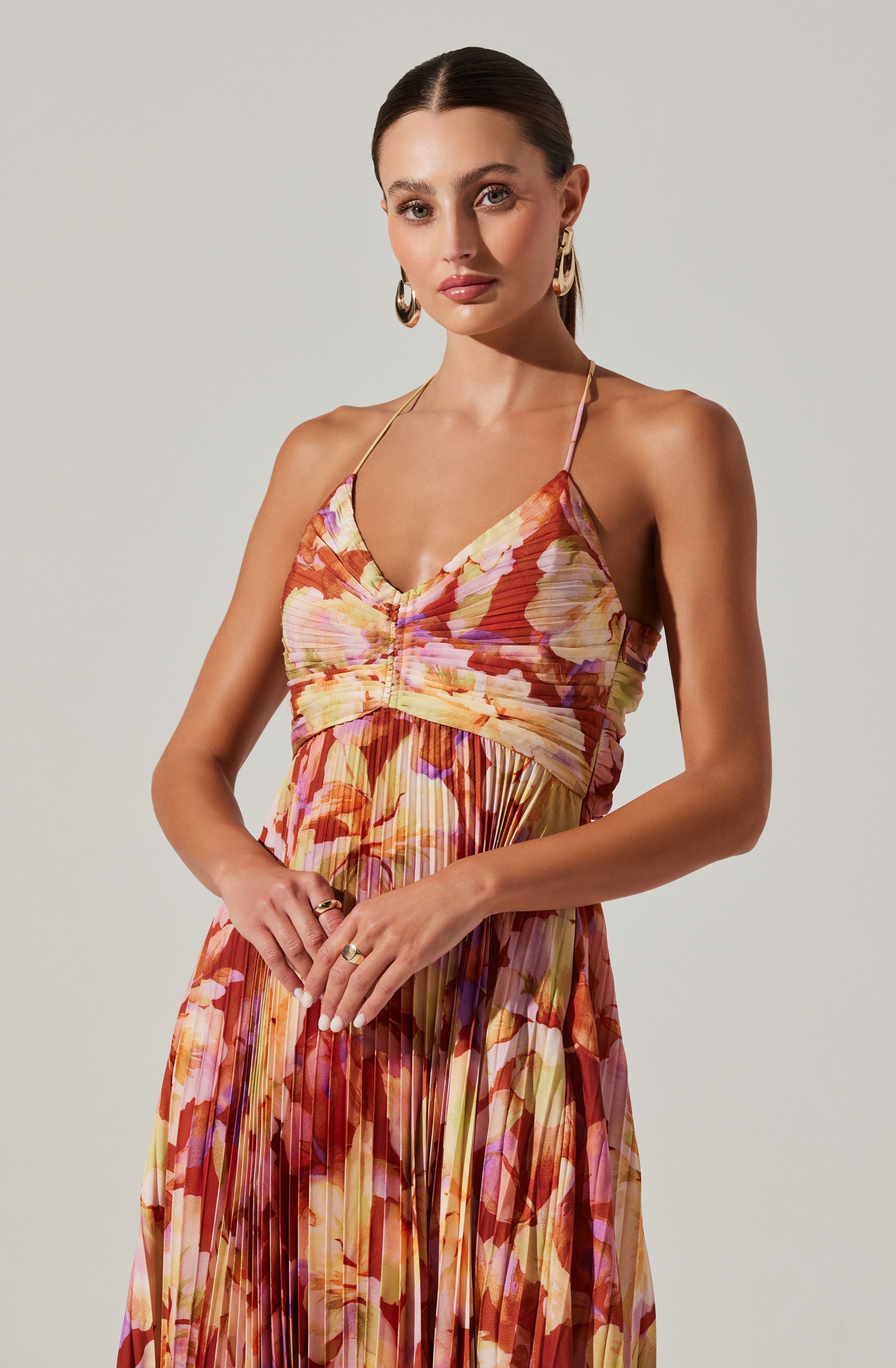 A woman is wearing a sleeveless, floral-printed dress with a fitted bodice and a flowing, pleated skirt. The dress features thin spaghetti straps and a gathered detail at the front of the bust. The fabric displays a vibrant mix of warm colors including shades of orange, pink, yellow, and purple with floral patterns. She is accessorized with large, gold hoop earrings and several rings on her fingers.