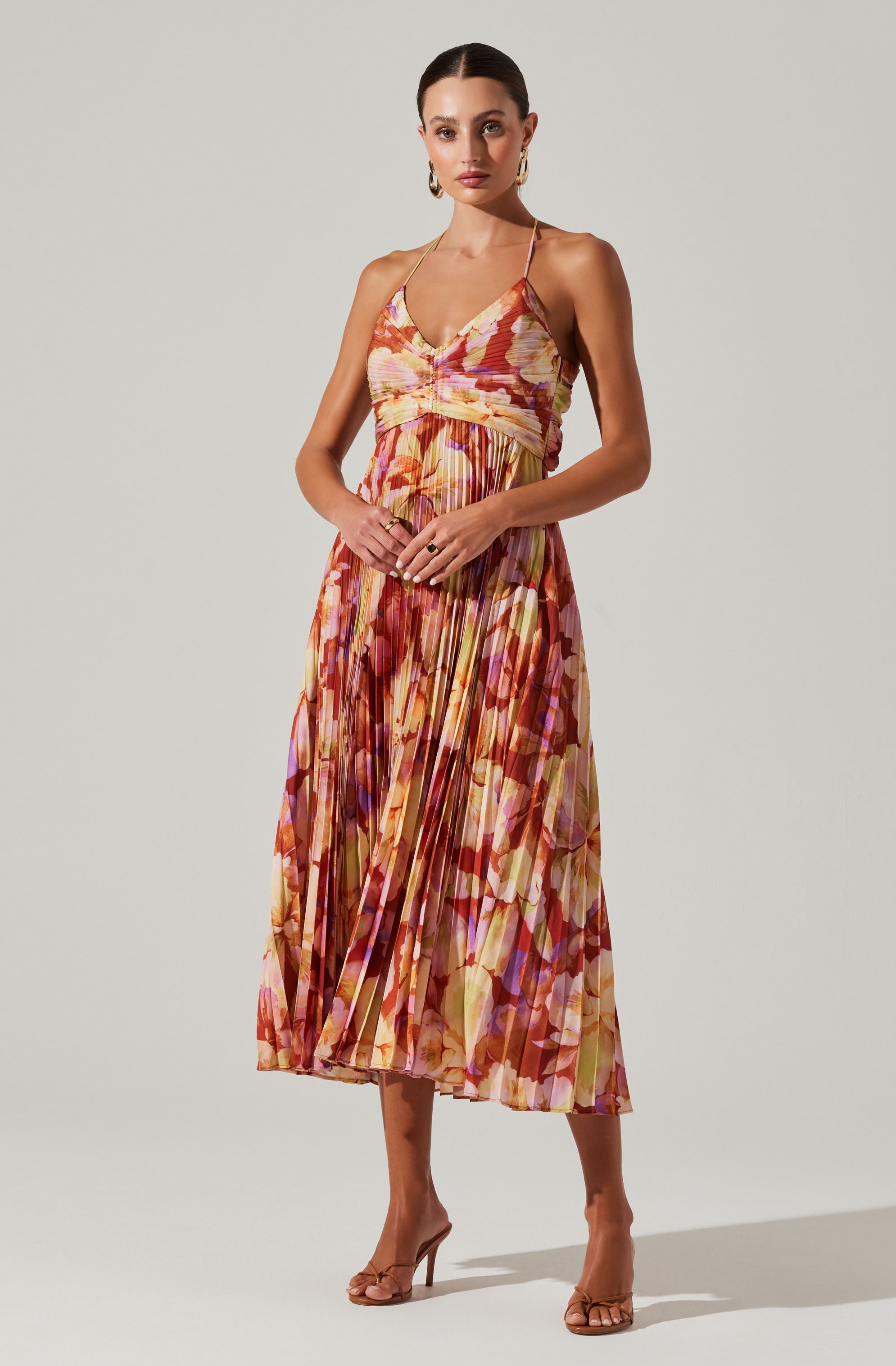 A woman is wearing a long, flowy dress with thin spaghetti straps. The dress features a vibrant, multicolored floral pattern with shades of pink, orange, yellow, and purple, and has a pleated skirt that reaches mid-calf. She is accessorized with large gold hoop earrings, a couple of rings, and beige high-heeled sandals.