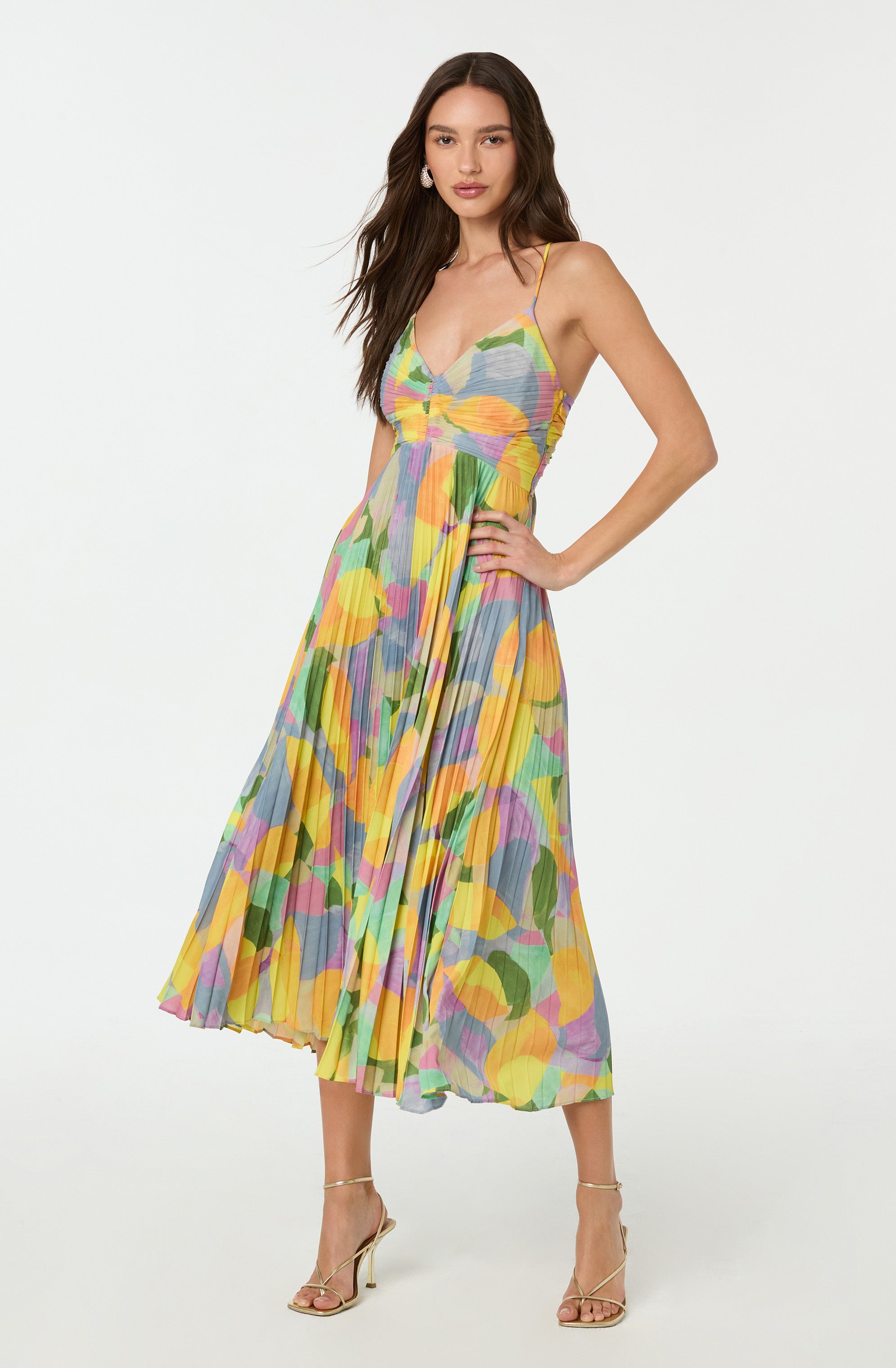 A woman is wearing a colorful, sleeveless, ankle-length dress with thin spaghetti straps. The dress features a vibrant abstract pattern with shades of yellow, green, pink, purple, blue, and orange, and has a pleated skirt. She is also wearing beige high-heeled sandals.